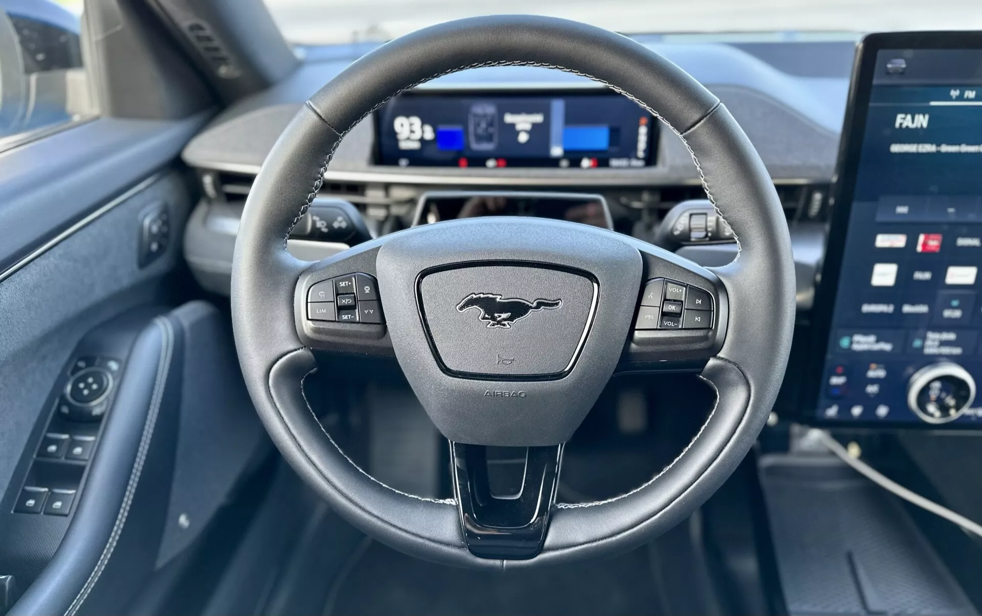 Vista interior de um Ford Mustang-Mach-E 2020 pela perspectiva do condutor, destacando um volante com três raios e controles, um painel de instrumentos digital e uma grande tela de infotainment sensível ao toque