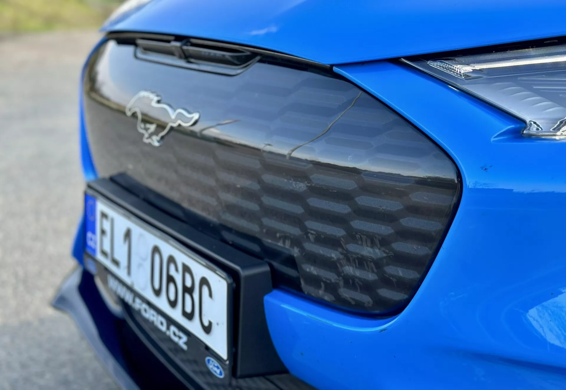 Vista aproximada da seção traseira de um Ford Mustang Mach-E 2020 azul, exibindo o emblema de cavalo cromado, spoiler traseiro em fibra de carbono, nicho integrado para placa de matrícula e visão parcial das luzes traseiras LED, acentuando as características aerodinâmicas e de desempenho do carro.