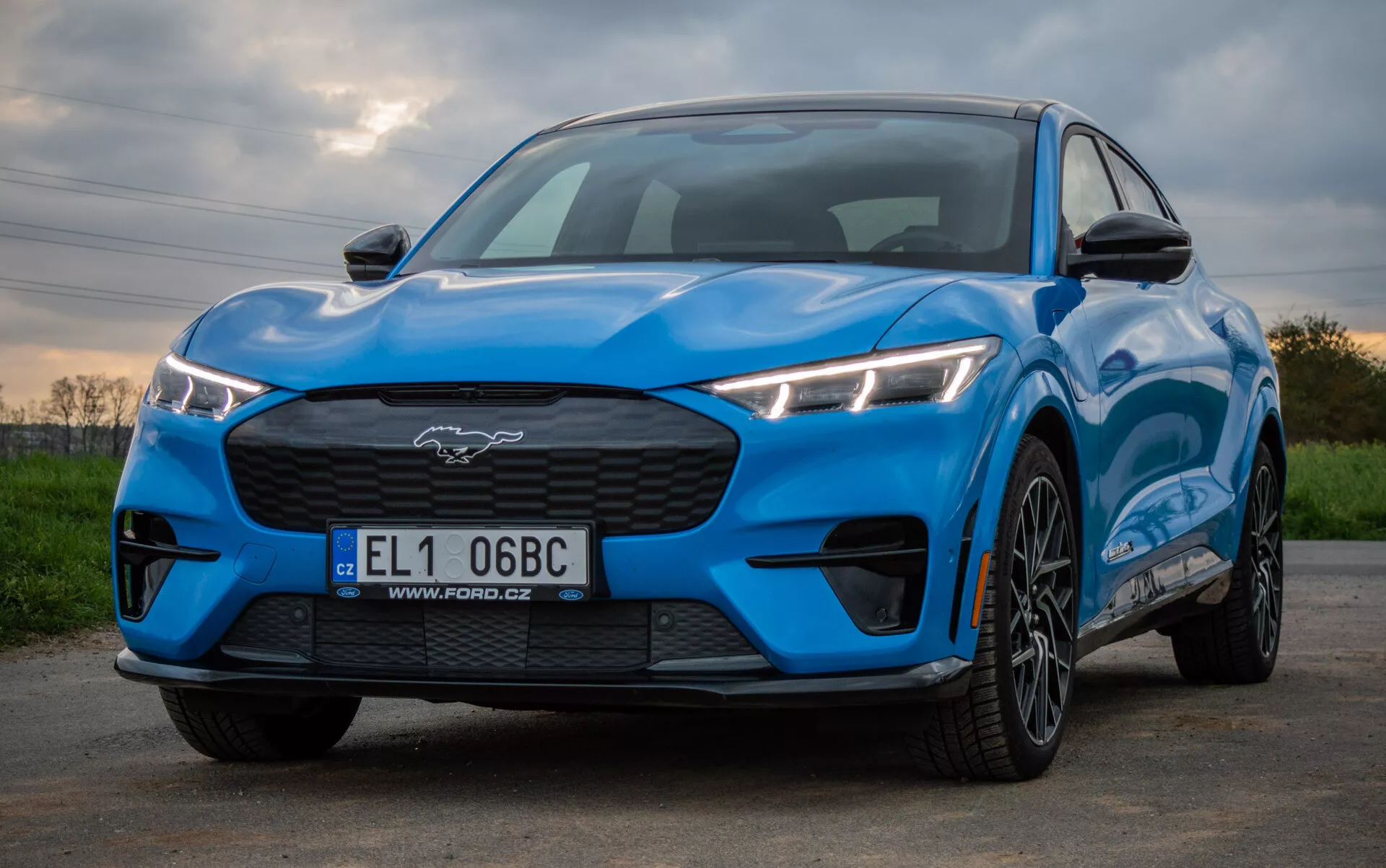 Ford Mustang Mach-E 2020 em azul brilhante exibindo um design esportivo e aerodinâmico de uma perspectiva frontal e lateral baixa, destacando faróis LED angulares, grelha fechada, capô contornado e rodas de liga leve de múltiplos raios contra um fundo natural.
