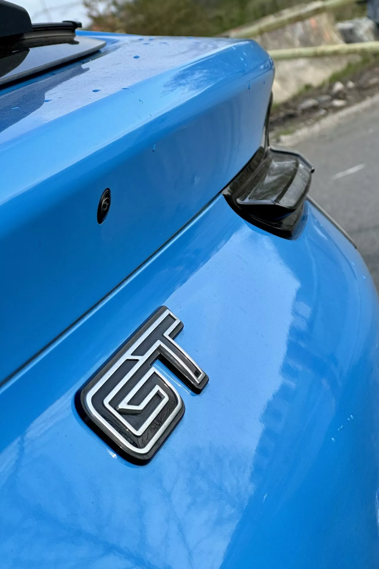 Vista detalhada da seção traseira de um Ford Mustang Mach-E azul 2020 focando no emblema GT metálico, porta-malas curvado e spoiler traseiro aerodinâmico com uma tampa de extremidade preta, contra um fundo levemente desfocado.