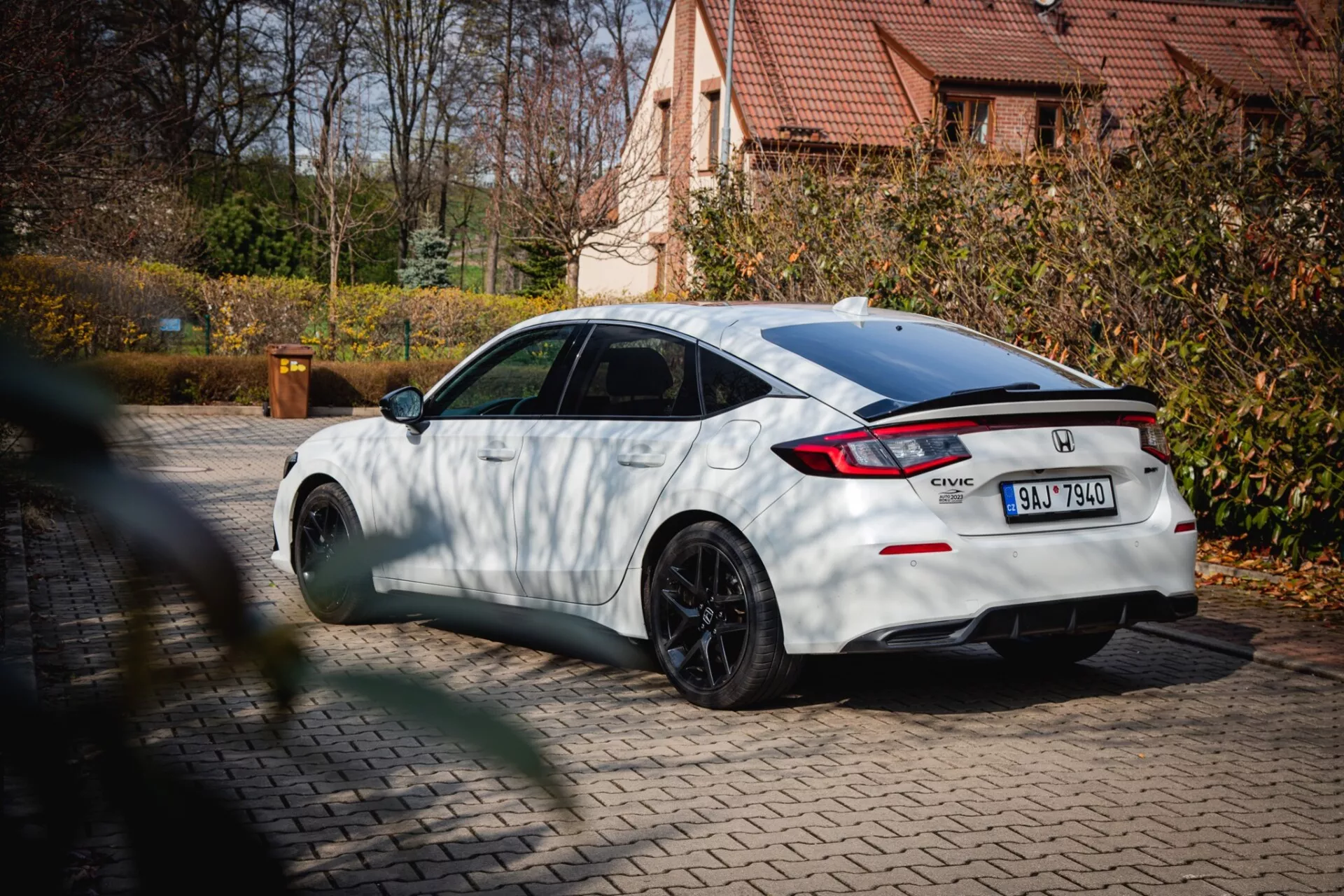 Honda Civic branco de 2022 com rodas de multi-raios pretas e spoiler traseiro, estacionado em pavimento com folhagem de outono e casa residencial ao fundo