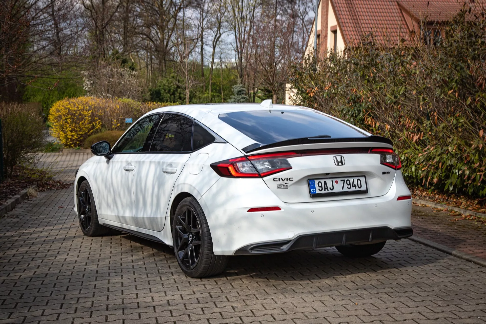 Honda Civic branco de 2022 mostrando perfil traseiro e lateral parcial em uma entrada de tijolos, com linha de telhado inclinada, spoiler integrado, luz de freio LED, luzes traseiras distintas e para-choque esportivo com difusor