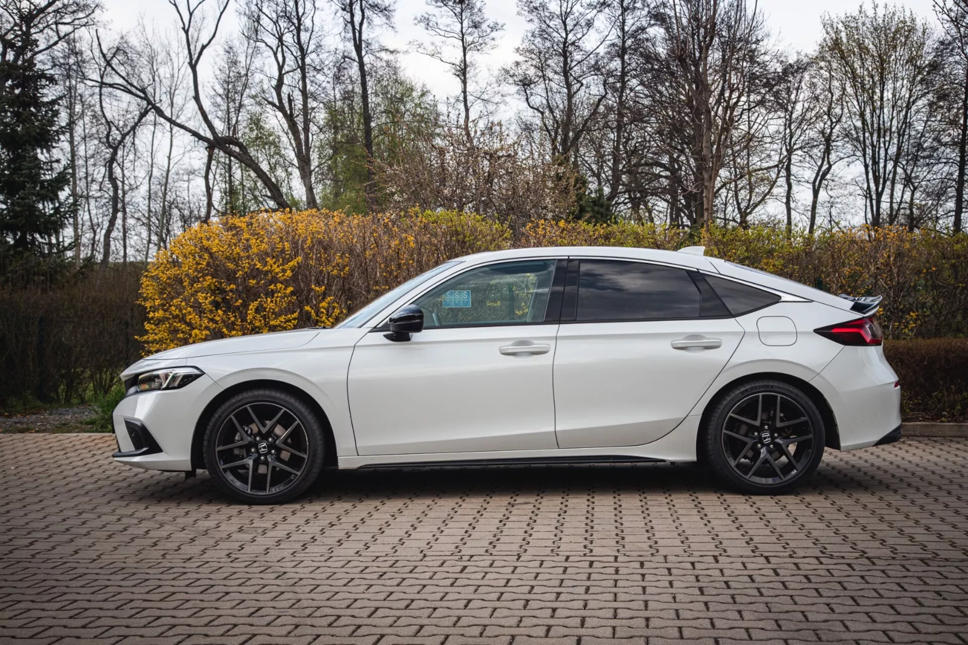 Sedan Honda Civic branco de quatro portas, modelo 2022, estacionado em uma área pavimentada com tijolos intertravados, exibindo um perfil lateral com design esportivo e faróis modernos de LED, capturado de um ângulo ligeiramente elevado para destacar a vista completa do lado incluindo janelas com película, rodas de liga leve esportivas e características tecnológicas como setas integradas nos retrovisores laterais.