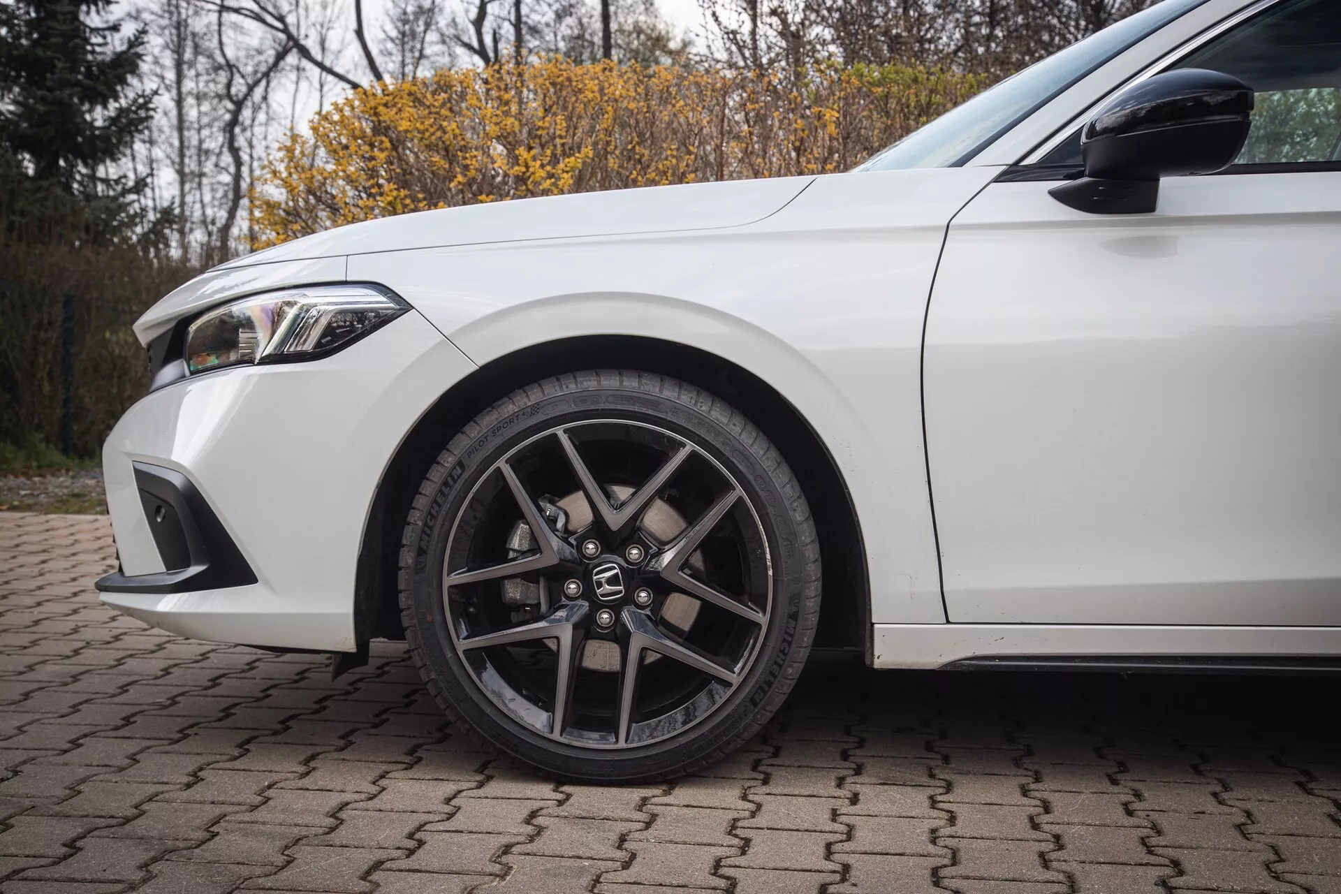 Honda Civic 2022 branco, exibindo roda dianteira esquerda com design de liga de múltiplos raios e pneu de perfil baixo, juntamente com detalhes do para-lama e parte do para-choque dianteiro sobre uma superfície de tijolo