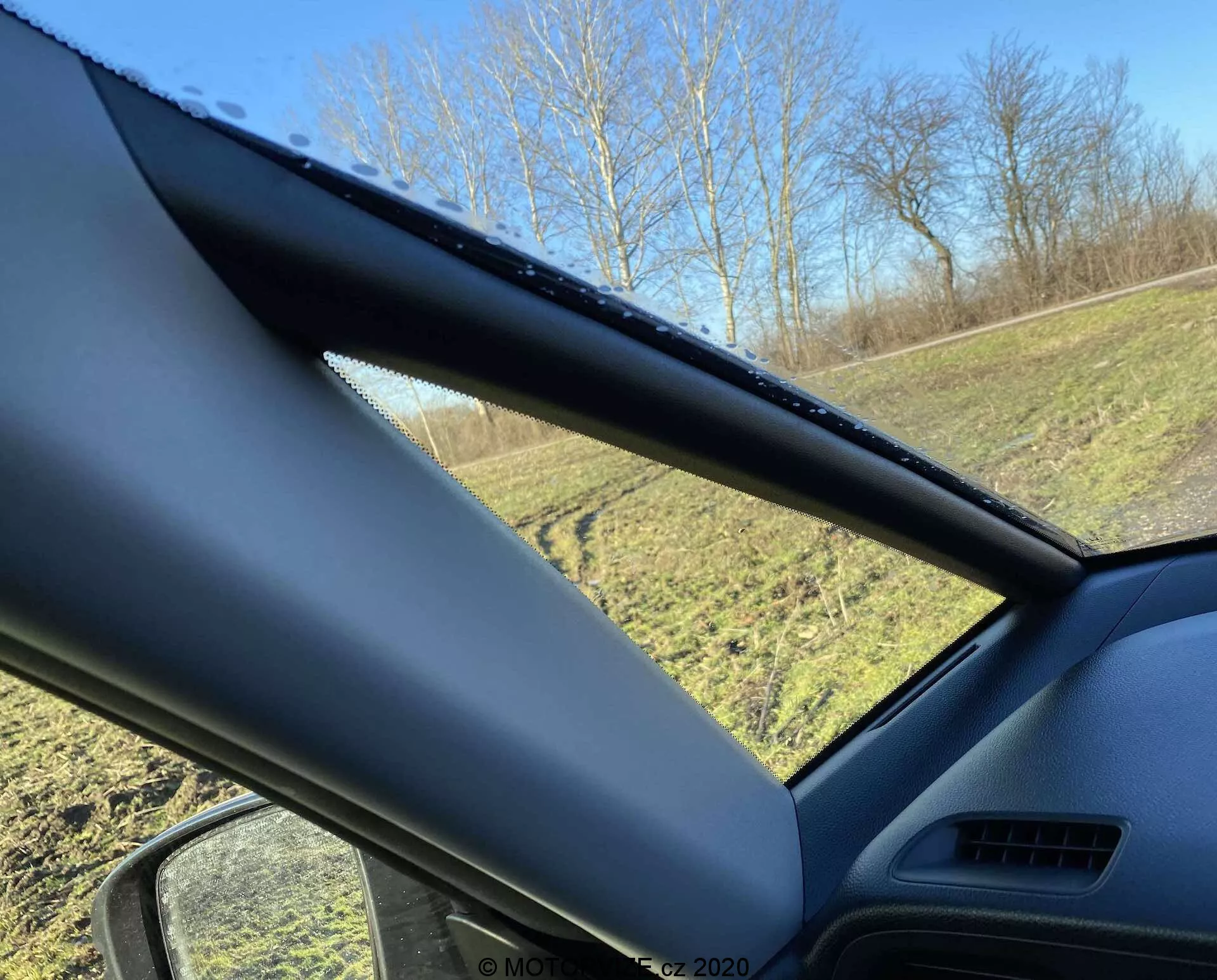 Vista interna de um Honda Jazz Crosstar 2020 olhando para a janela do lado do motorista, focando no pilar A. O pilar A é coberto com um material texturizado não metálico e obstrui parcialmente a vista. A janela lateral mostra gotas de água, indicando chuva recente ou condensação, com uma área gramada e árvores visíveis do lado de fora sob um céu azul claro.