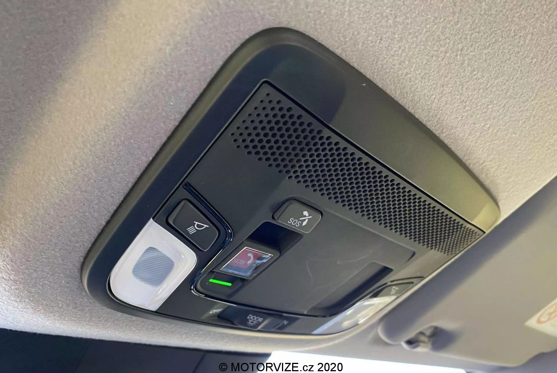 Vista em close-up do console superior do Honda Jazz 2020 (Jazz Crosstar). A imagem revela uma série de botões funcionais, incluindo controles de luz interna, um possível botão de destravamento de porta, uma área ventilada com microfone para chamadas mãos-livres, um home link com botões programáveis para abertura de portas de garagem e um possível compartimento para óculos de sol ou fecho de armazenamento. O console é capturado à luz do dia com foco claro nos botões texturizados, destacando a tecnologia avançada e o design interior do veículo.