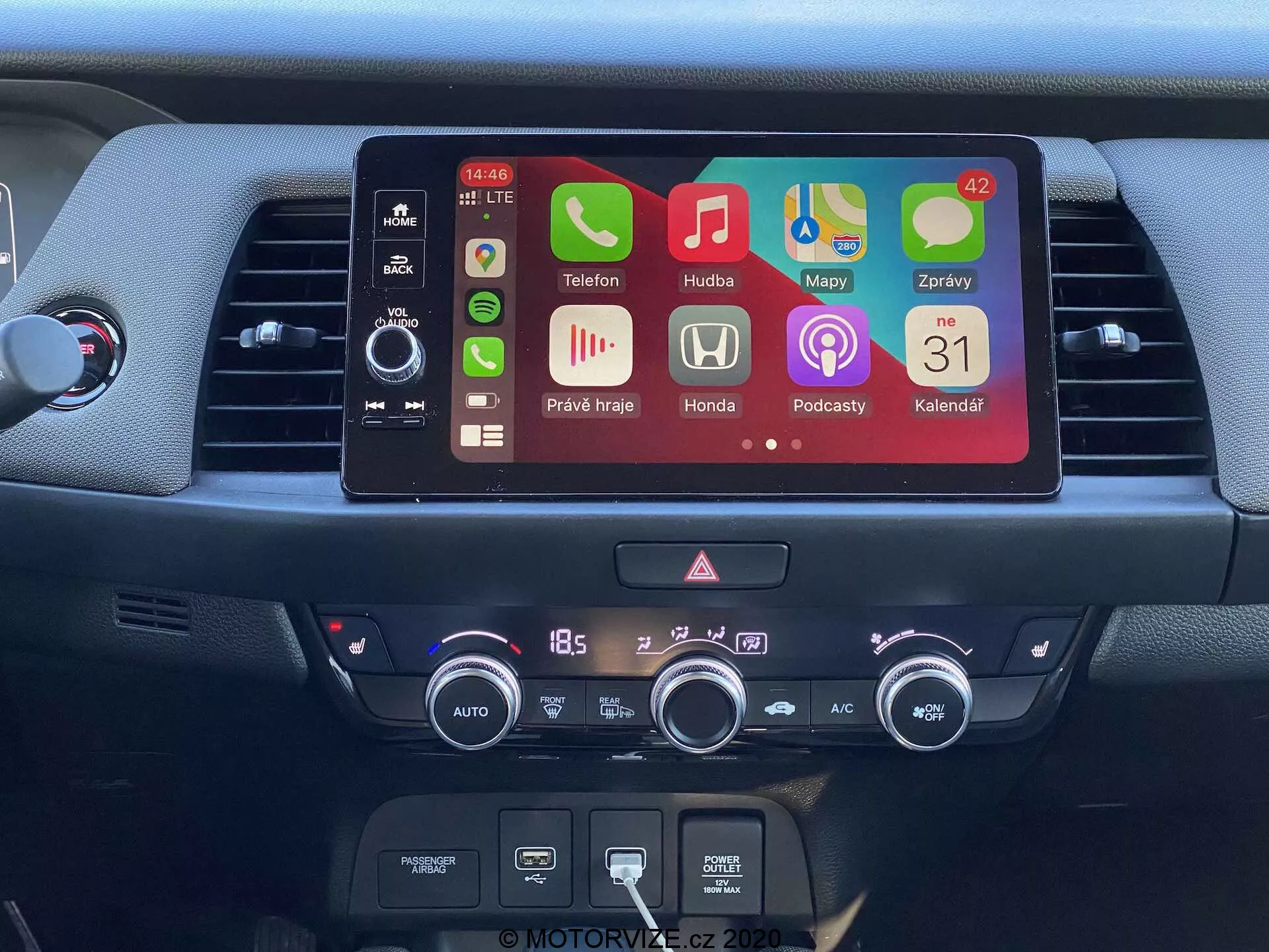 Visão detalhada do console central do Honda Jazz 2020, apresentando uma tela moderna de infotainment com ícones de aplicativos coloridos incluindo telefone, música, navegação, configurações do carro, podcasts e calendário. Abaixo da tela encontram-se controles físicos do sistema de áudio, uma ranhura para CD e um sistema de controle climático digital ajustado para 18,5 graus Celsius. A imagem destaca recursos avançados como conectividade Bluetooth, integração de smartphones e controle climático automático.