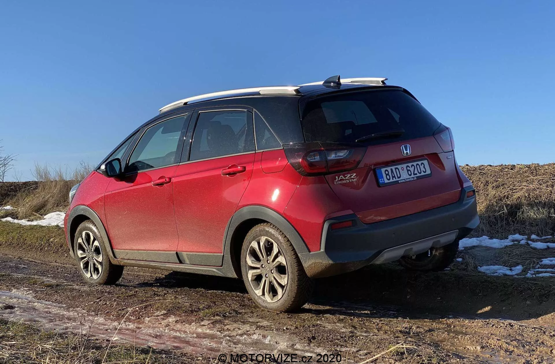 Um SUV Honda Jazz Crosstar 2020 vermelho visto de três quartos traseiros, mostrando detalhes como emblemas de marca e modelo, luzes traseiras em LED, para-choques traseiro com arranhões e sensores, rodas de liga leve, pneus para todas as estações, trilhos de teto, antena de barbatana de tubarão, vidros escurecidos com linhas de desembaçamento e placa de licença centralizada. A abertura do porta-malas tipo hatchback e manchas de neve no chão com um campo seco ao fundo sugerem um cenário de final de outono, inverno ou início da primavera sob um céu claro ao meio-dia ou no início da tarde.