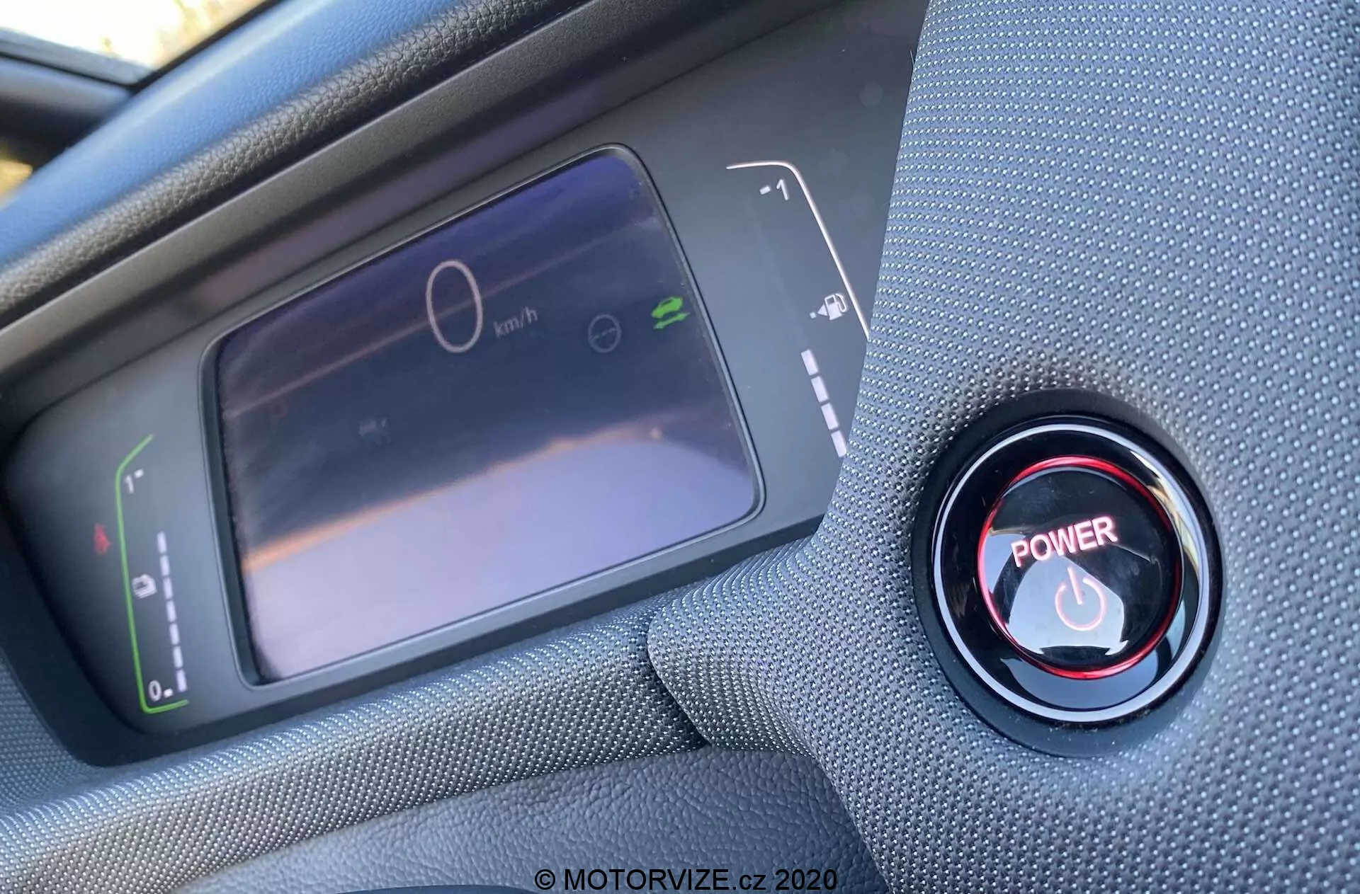 Vista interna em close-up a partir da perspectiva do motorista de um Honda Jazz Crosstar 2020, focando no display digital do painel de instrumentos e no botão de partida/parada do motor vermelho. O display LCD à esquerda mostra um velocímetro estacionário e um gráfico de uso de energia, enquanto à direita, o botão vermelho de partida/parada é cercado por um anel metálico. A foto foi tirada em um ângulo oblíquo de cima, com foco nítido no botão e foco suave no display.