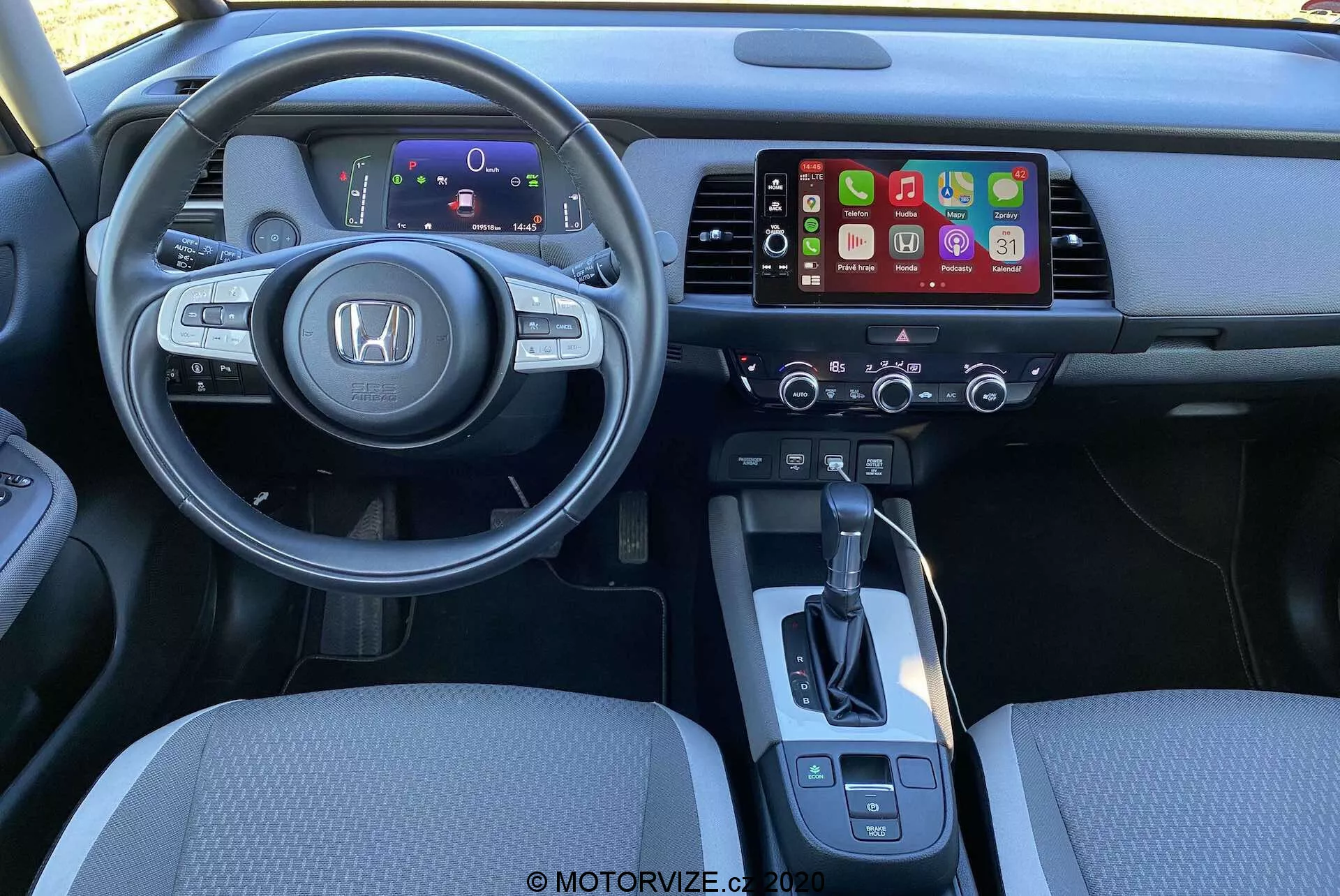 Vista interior do Honda Jazz Crosstar 2020 a partir da perspectiva do motorista. Apresenta um volante moderno e multifuncional com o logotipo da Honda em destaque, completo com controles de áudio, controle de cruzeiro e comando de voz. Atrás do volante há um painel de instrumentos digital que mostra informações essenciais. O console central inclui um sistema de infotainment que provavelmente suporta Apple CarPlay ou Android Auto, com controles de clima abaixo. A alavanca de câmbio é simples, com controles adicionais para o freio de mão eletrônico e seleção de modo de condução. A iluminação e o ângulo destacam o conforto e o design ergonômico.