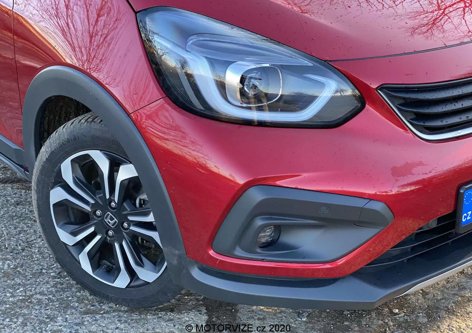 Close-up da esquina frontal de um Honda Jazz 2020 (Jazz Crosstar) vermelho, focando no moderno farol de LED, detalhe minimalista da grade, para-choque dianteiro bicolor com farol de neblina e roda de liga leve de cinco raios dinâmica.