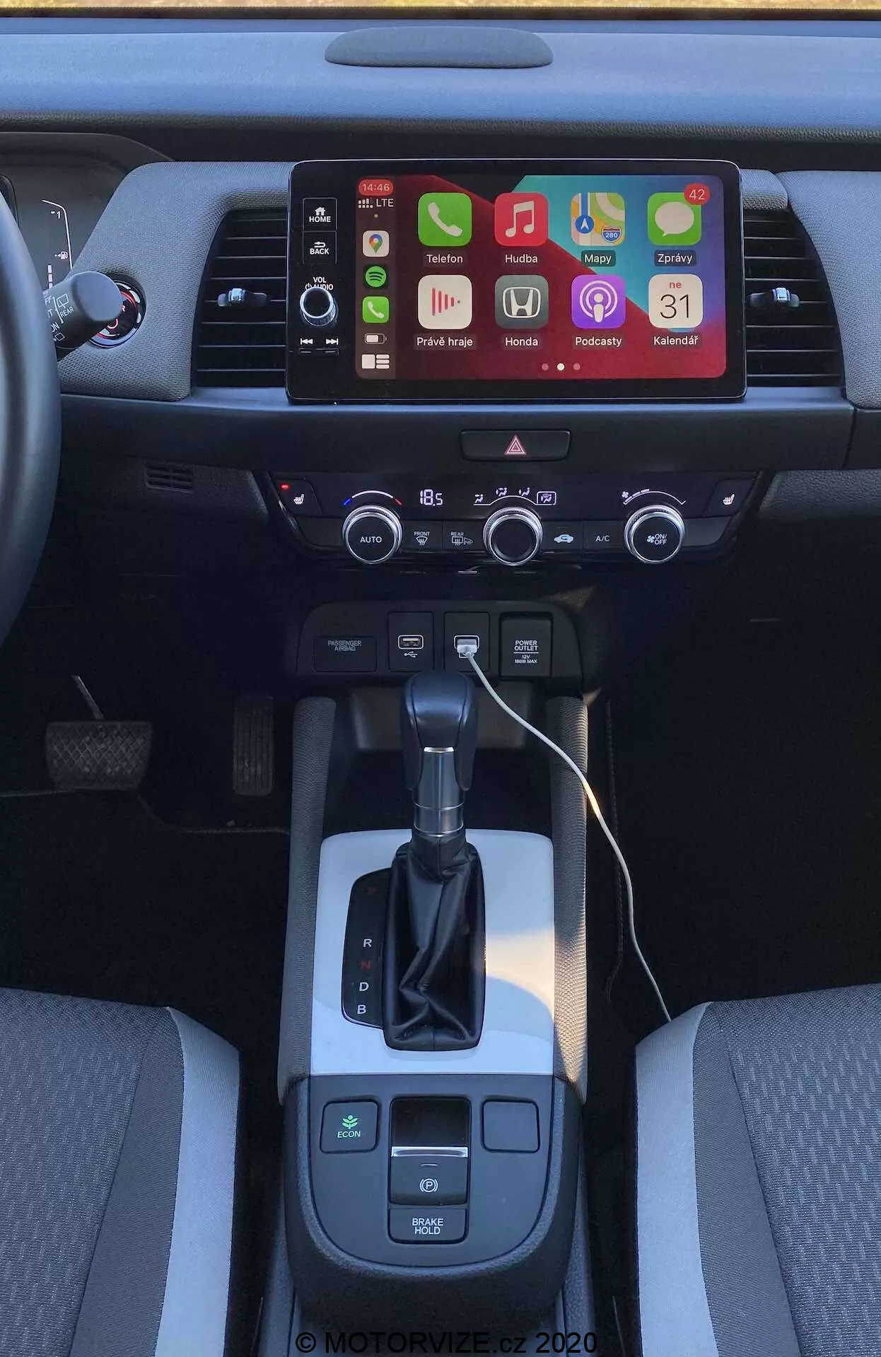 Vista interior da consola central do Honda Jazz Crosstar 2020 a partir de um ângulo ligeiramente elevado para trás. Foco na área da alavanca de câmbio, seletor de transmissão automática e ecrã tátil de info-entretenimento com vários ícones de aplicativos, sugerindo opções avançadas de conectividade como Apple CarPlay e Android Auto. Abaixo do ecrã, há aberturas de ventilação e um painel de controlo climático de dupla zona com botões e mostradores físicos. A alavanca de câmbio é flanqueada por seletor de modo de condução e botões de travão de estacionamento eletrónico, com um espaço de armazenamento aberto à direita, indicando portas de carregamento. Os bancos dianteiros com estofos de duas cores destacam o design interior e o conforto.