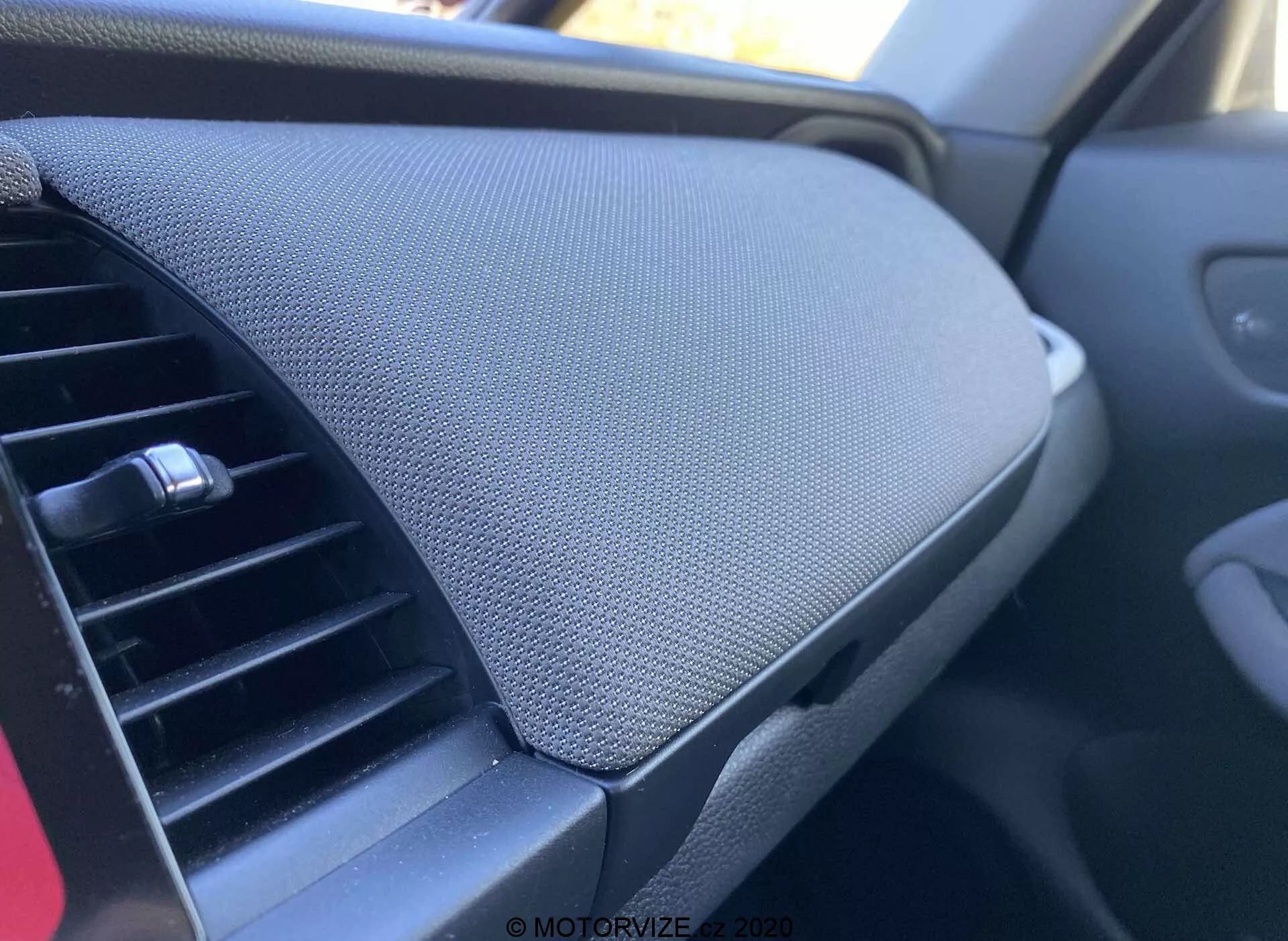 Vista aproximada do interior de um veículo Honda Jazz 2020 (Jazz Crosstar), focada no painel. Tirada da perspectiva do passageiro, a imagem mostra o ventilador de ar condicionado à esquerda, com design horizontal ajustável. Dominando o lado direito da imagem está o compartimento de armazenamento do painel com tampa fechada, caracterizado por uma superfície texturizada, possivelmente de malha ou tecido. O compartimento parece integrado sem uma alça visível, sugerindo um mecanismo de abertura por pressão. O esquema de cores geral é escuro, implicando uma estética neutra destinada a reduzir as distrações.