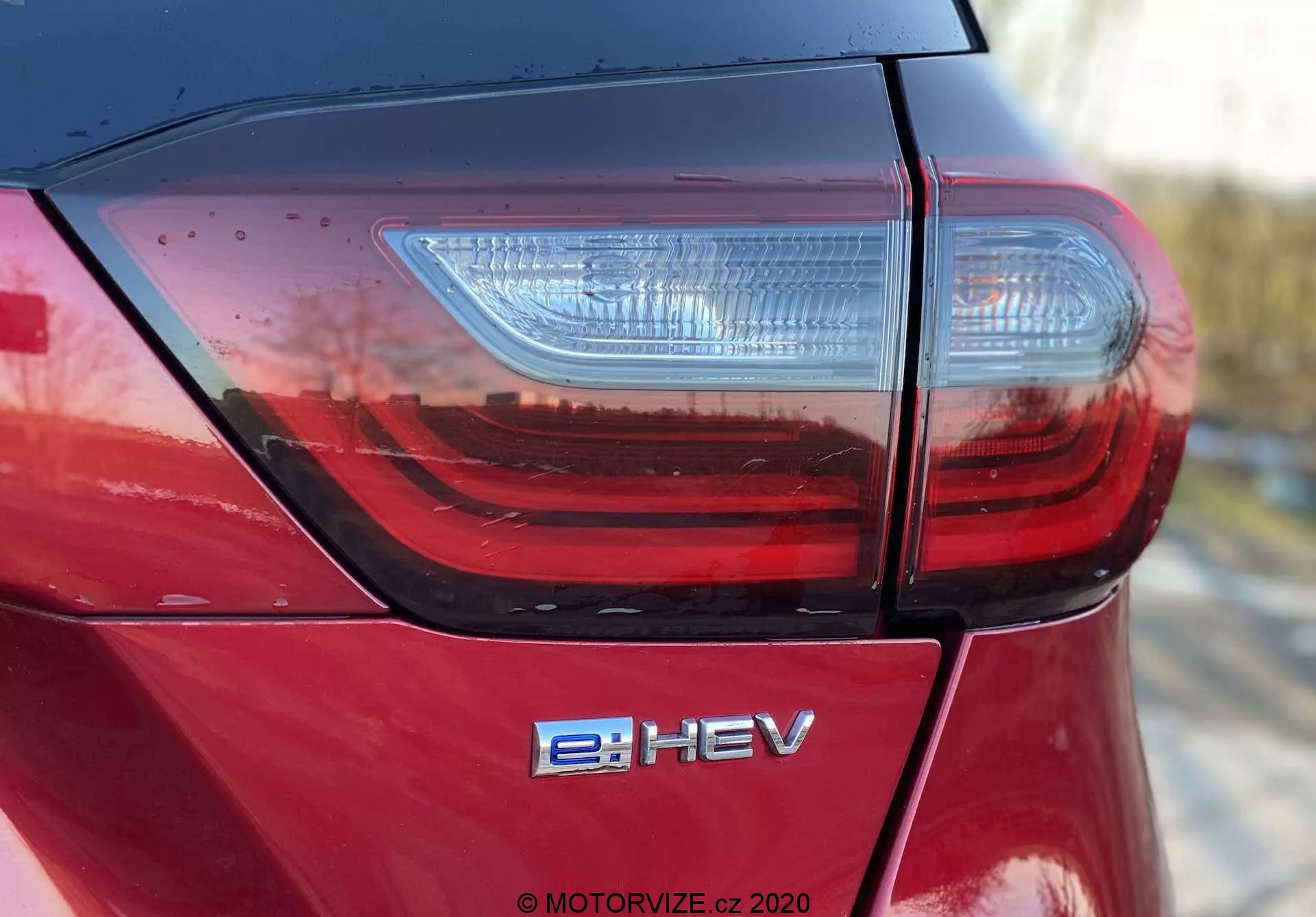 Foto detalhado de ângulo alto da seção traseira de um Honda Jazz Crosstar 2020 capturando a tampa do porta-malas, o moderno conjunto de luzes traseiras LED e o emblema de veículo elétrico híbrido e:HEV. Destaca o acabamento de pintura metálica vermelha de alto brilho e a qualidade de construção superior com emendas de painéis bem ajustadas, indicadas pelas linhas limpas e reflexos na luz natural.