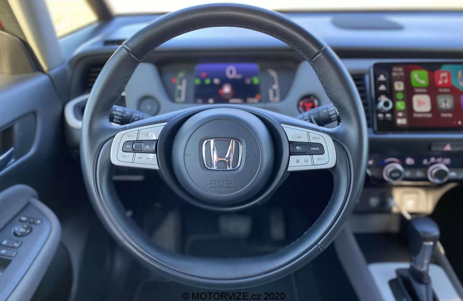 Vista do interior do Honda Jazz Crosstar 2020 a partir do assento do motorista, com destaque para um volante com botões de controle de áudio, controle por voz e controle de cruzeiro. O emblema da Honda é exibido de maneira proeminente no centro. Atrás do volante, um painel de instrumentos digital exibe vários indicadores e informações. À direita, um sistema de infotainment com tela sensível ao toque e compatibilidade com Apple CarPlay está visível, juntamente com controles de clima ligeiramente desfocados. A imagem, capturada de uma perspectiva em primeira pessoa, destaca o design ergonômico e a acessibilidade da tecnologia do veículo.