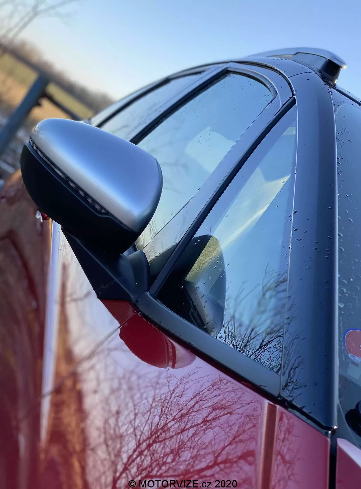Vista lateral aproximada do Honda Jazz 2020 (Jazz Crosstar) destacando o espelho retrovisor do lado do motorista com indicadores de seta integrados. A imagem, tirada de um ângulo baixo próximo à frente do carro, capta a carroceria brilhante, possivelmente vermelha, com gotículas de água, sugerindo chuva recente ou orvalho matinal. O espelho com carcaça escura e o design aerodinâmico do carro são destacados, enfatizando a qualidade do acabamento e do exterior.
