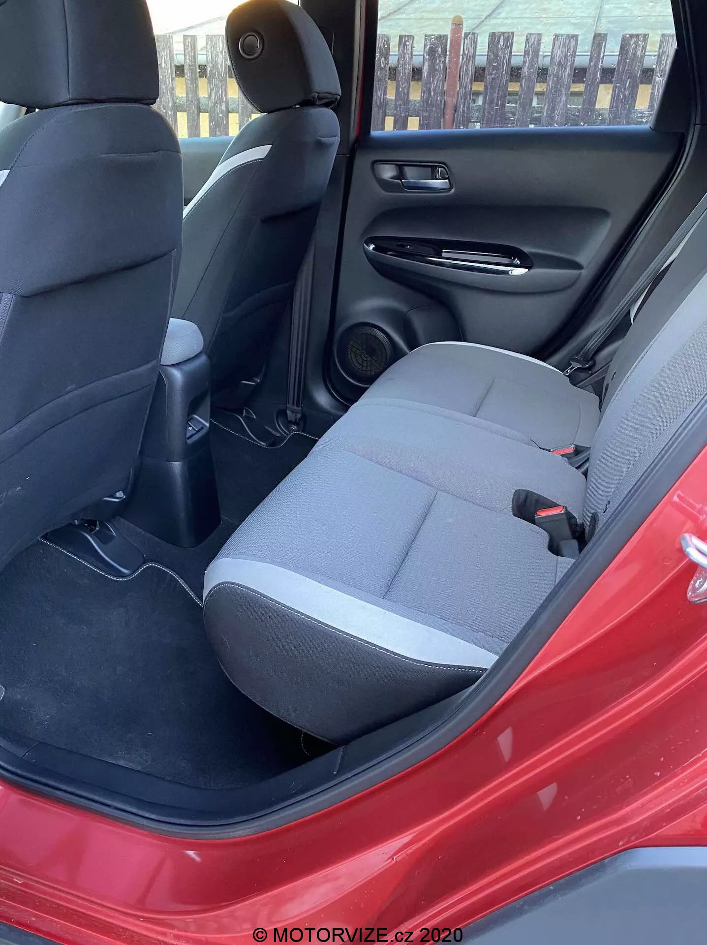Vista do interior do Honda Jazz Crosstar 2020 pela porta traseira do lado do condutor aberta, destacando o banco traseiro do passageiro em tecido escuro, cinto de segurança, consola central traseira com ventiladores, altifalante na porta traseira, painel interno preto com bolsa de arrumação, piso tapetado e a cor vermelha marcante do exterior do carro. A luz natural do dia ilumina a cena.