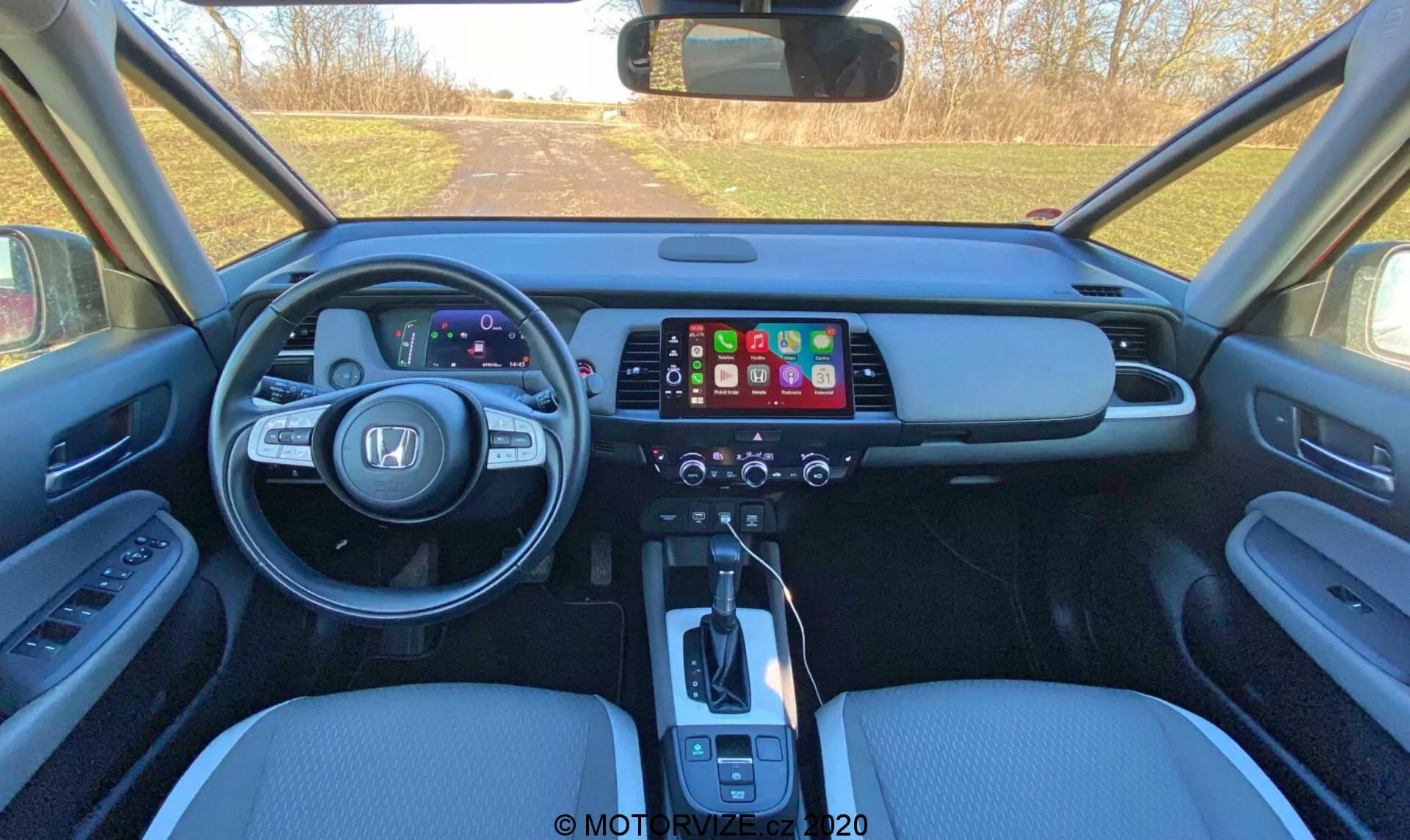 Vista interior de um Honda Jazz 2020 (Jazz Crosstar) moderno a partir da área dos bancos traseiros, mostrando o painel frontal com painel de instrumentos digital, grande ecrã tátil de infotainment, painel de controle de clima, alavanca de câmbio automática, assentos em couro e controles do painel da porta.