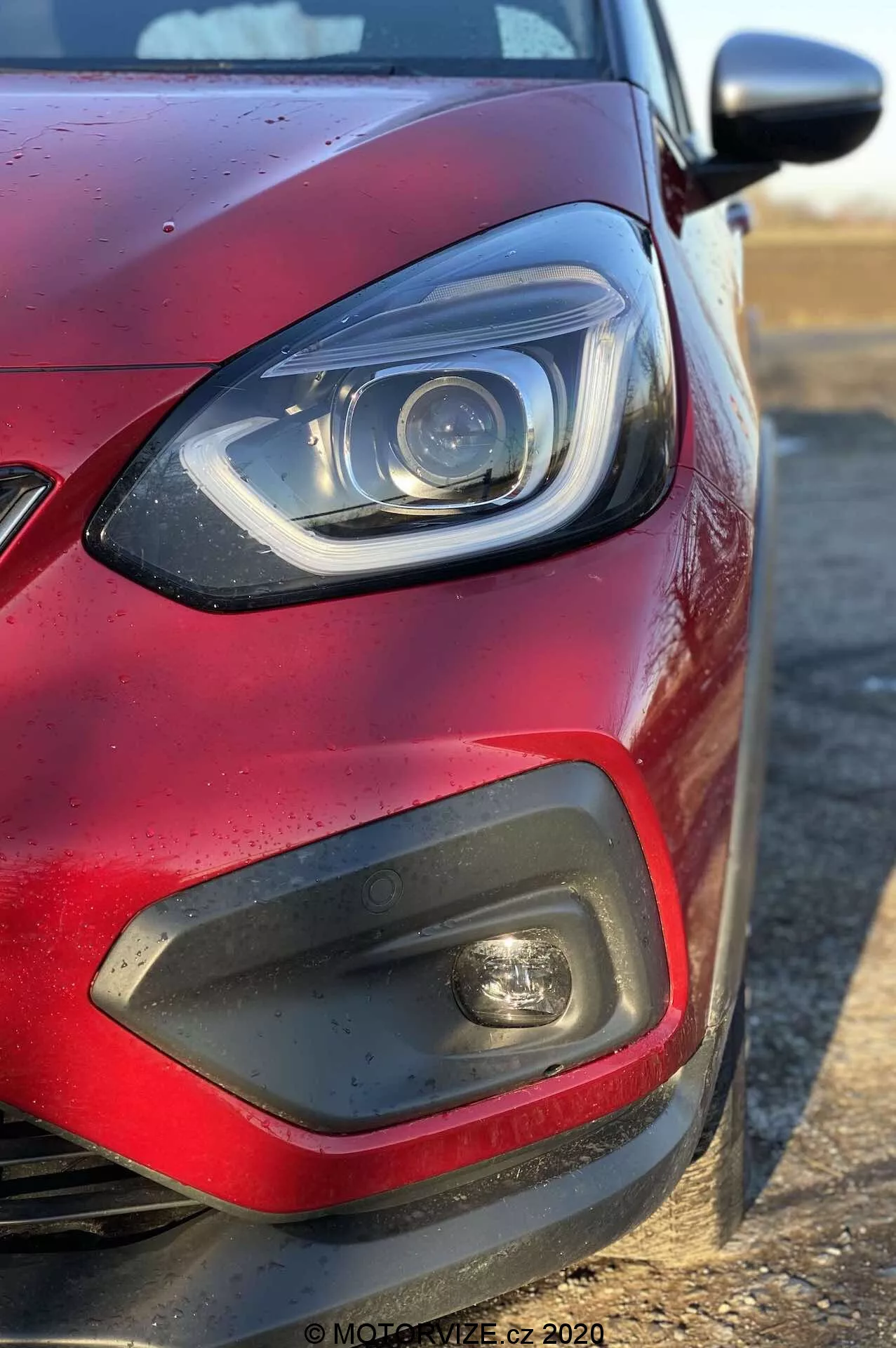 Close-up do quadrante dianteiro esquerdo de um Honda Jazz 2020 (Jazz-Crosstar) vermelho de um ângulo lateral, focando no farol com elementos LED ou xenon e uma barra DRL curva, um inserto de plástico preto com luz de nevoeiro, uma parte da grade, pintura metálica vermelha com gotas de água e um espelho lateral na cor da carroceria estendendo-se da direita.
