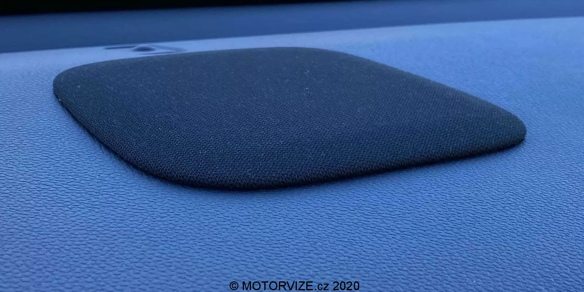 Fotografia em close-up de uma grade de alto-falante de carro embutida em um painel de instrumentos ou painel de porta azul escuro ou preto, possivelmente de um Honda Jazz 2020. A grade do alto-falante é feita de um material texturizado semelhante a tecido e possui a forma de trapezoide truncado ou quadrilátero irregular com um lado ligeiramente convexo. O componente está ligeiramente elevado, projetando uma sombra que enfatiza seu perfil e textura. A foto foi tirada com um ângulo lateral usando luz natural difusa, destacando a textura granulada da superfície circundante e contém uma marca d'água que diz '© MOTORVIZE.cz 2020.'