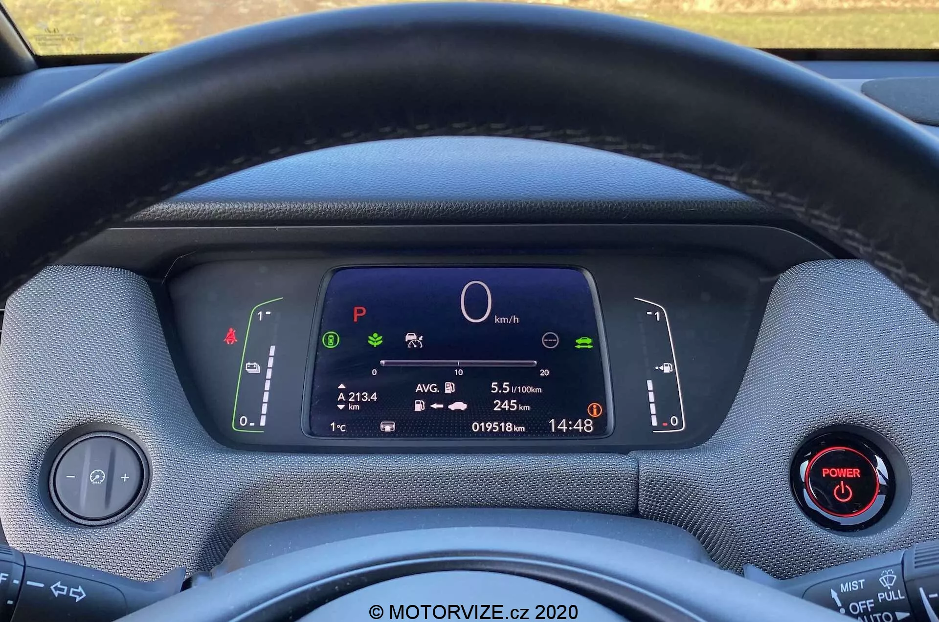 Close-up do interior do Honda Jazz Crosstar 2020 visto do assento do motorista, mostrando o volante e o painel de instrumentos totalmente digital. O volante envolto em tecido obstrui parcialmente a visão do display digital, que apresenta símbolos do sistema do carro, um gráfico circular central, informações de viagem, odômetro e relógio. Duas alavancas atrás do volante controlam as setas, faróis e limpadores de para-brisa. Um painel de controle para os faróis e um botão de ligar com um anel vermelho são visíveis à esquerda e à direita, respectivamente. Os materiais do painel parecem texturizados, destacando um design ecológico. A imagem enfatiza a tecnologia moderna, instrumentação digital e possíveis características de veículos híbridos ou elétricos.