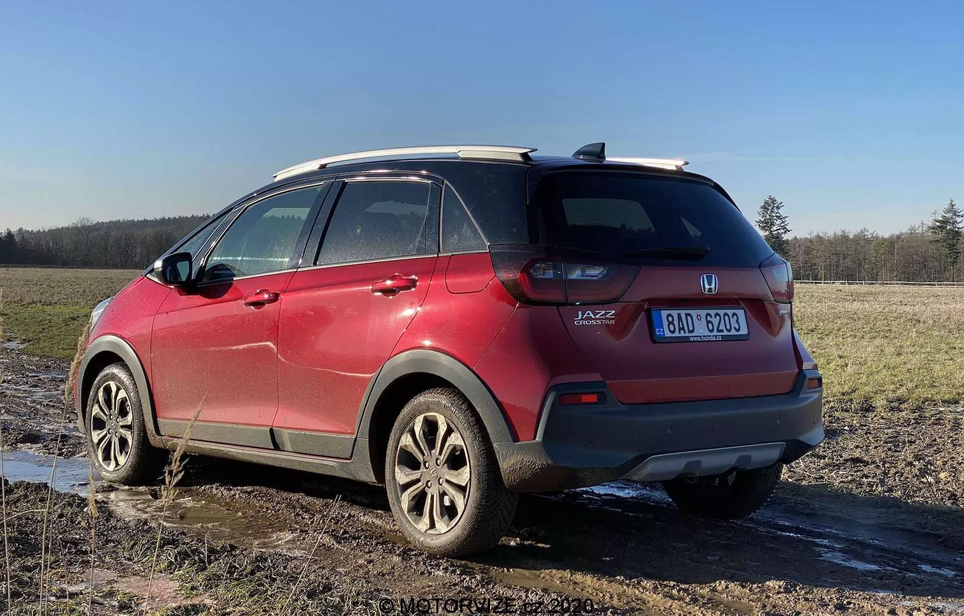 Vista traseira de três quartos de um Honda Jazz Crosstar 2020 vermelho, destacando suas características de SUV subcompacto crossover. A imagem capturada de um ângulo baixo destaca a altura e a distância ao solo do veículo, com clusters de luz traseira visíveis, insígnia 'JAZZ', rodas de liga leve, sistema de trilhos de teto, vidros traseiros escurecidos, respingos de lama e uma placa de licença visível, estacionado em um campo lamacento.