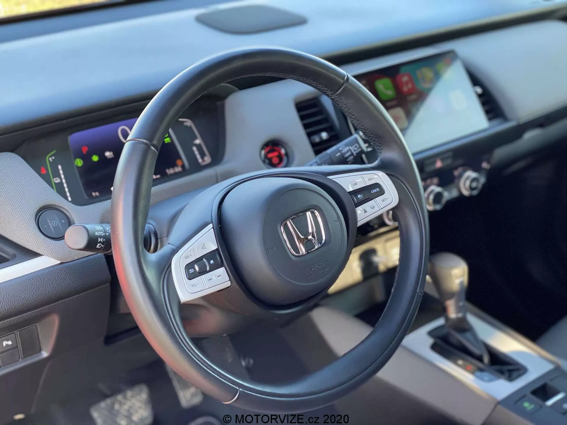 Close-up do interior do Honda Jazz Crosstar 2020, capturado a partir da perspectiva do banco do motorista, destacando o lado esquerdo do volante de três raios revestido em couro com controles multifuncionais, e o painel de instrumentos com cluster digital e sistema de infotainment com tela sensível ao toque. A imagem inclui controles de HVAC e uma alavanca de câmbio automática revestida em couro na consola central, enfatizando um design moderno e bem equipado.