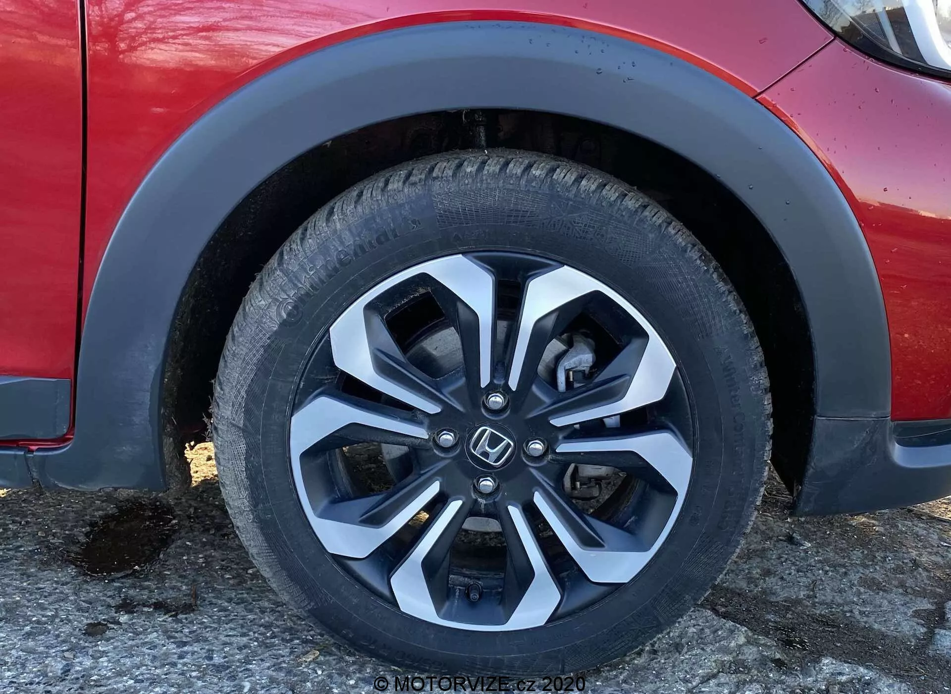 Imagem em close do pneu dianteiro esquerdo e da carroceria inferior do Honda Jazz Crosstar 2020, destacando a roda de liga leve moderna com múltiplos raios e acabamento em duas tonalidades (prata e preto), os detalhes do desenho da banda de rodagem e as informações da parede lateral do pneu. A imagem também mostra o arco do para-lama, parte do para-choque dianteiro e uma seção da porta lateral a partir de um ângulo ligeiramente baixo, enfatizando aspectos de design e tecnologia como a tecnologia dos pneus, design das rodas, composição do material, aerodinâmica e dicas sobre a suspensão do veículo.
