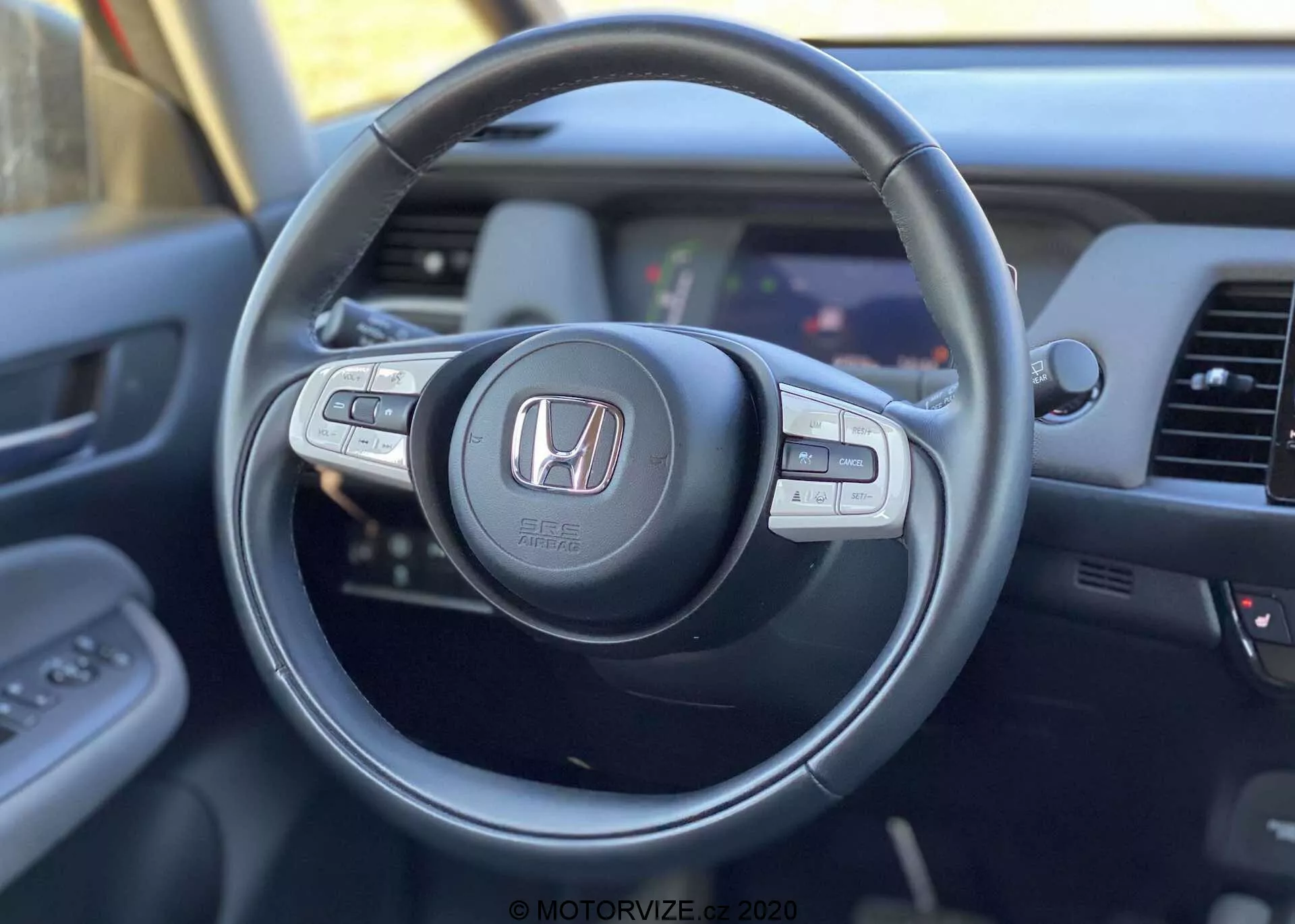 Vista interior de um Honda Jazz Crosstar 2020 a partir da perspetiva do condutor, focando no volante moderno com logótipo Honda, com botões de controlo multifuncional para áudio, controlo de velocidade de cruzeiro e chamadas em mãos-livres, com uma visão parcial do painel de instrumentos e do sistema de infotainment.