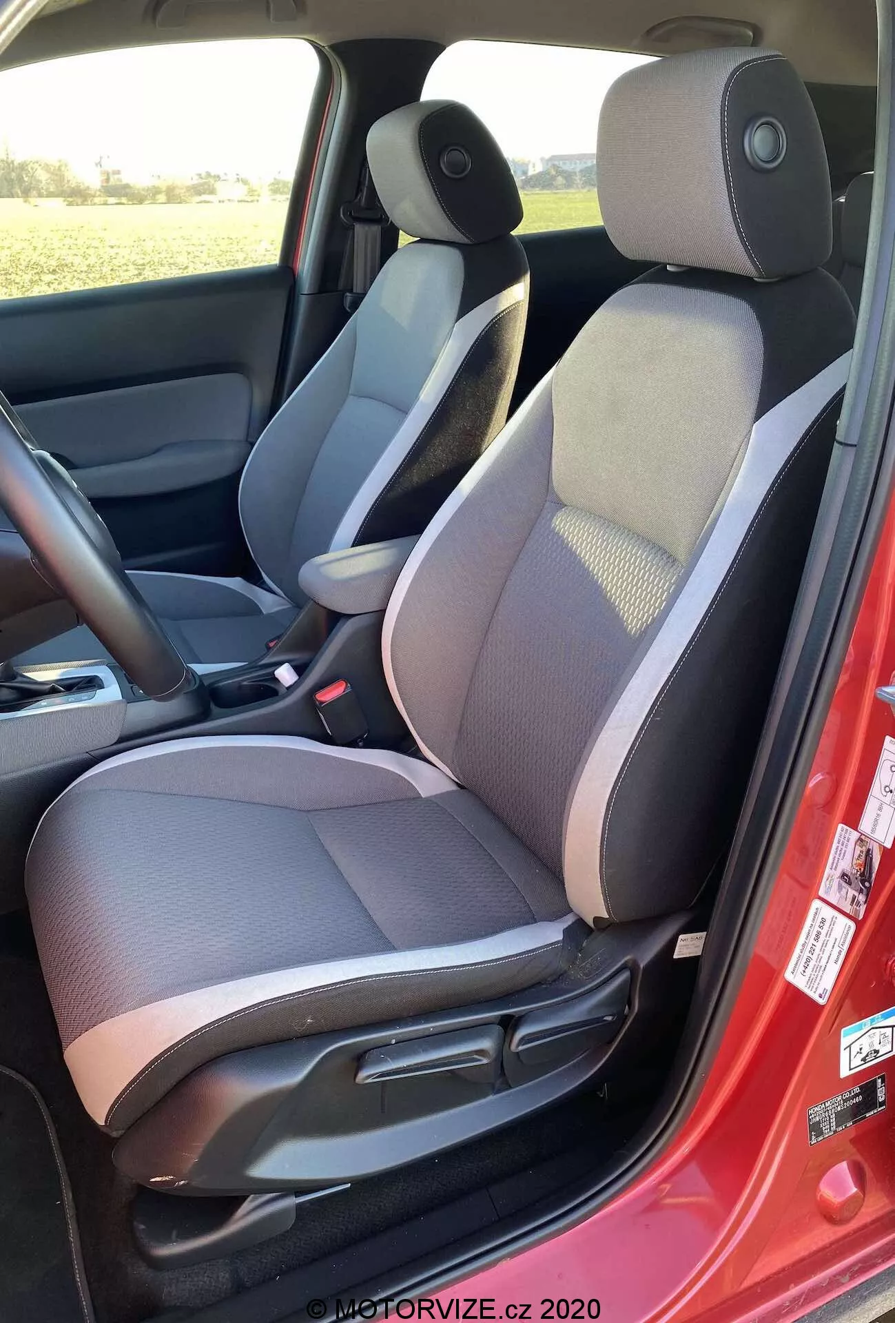 Vista interior do Honda Jazz Crosstar 2020 do lado esquerdo, destacando o banco do motorista em tecido de dois tons com seção central estampada, encosto de cabeça ajustável, controles manuais de ajuste do banco, cinto de segurança vermelho, alto-falante no painel da porta, tapete escuro, console central com apoio de braço, controle do espelho lateral e esquema de cores escuras e claras.