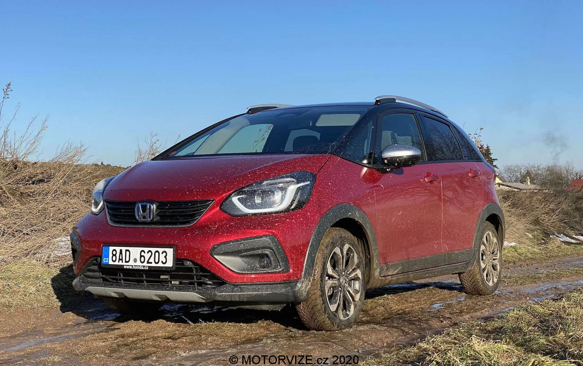 Uma vista de ângulo frontal de um Honda Jazz Crosstar 2020 vermelho, um SUV compacto crossover, capturado em terreno off-road com sujeira e lama visíveis. A imagem destaca a grelha frontal, os faróis elegantes, possivelmente com lâmpadas LED ou de projetor, os faróis de neblina integrados no para-choque e o capô ligeiramente inclinado. O veículo, com barras de teto, rodas cobertas de lama e espelhos laterais com sinalizadores integrados, sugere recursos modernos de segurança e equipamento tecnológico avançado. Ideal para categorias focadas em veículos, avaliações de carros, capacidades off-road, tecnologia automotiva e design.
