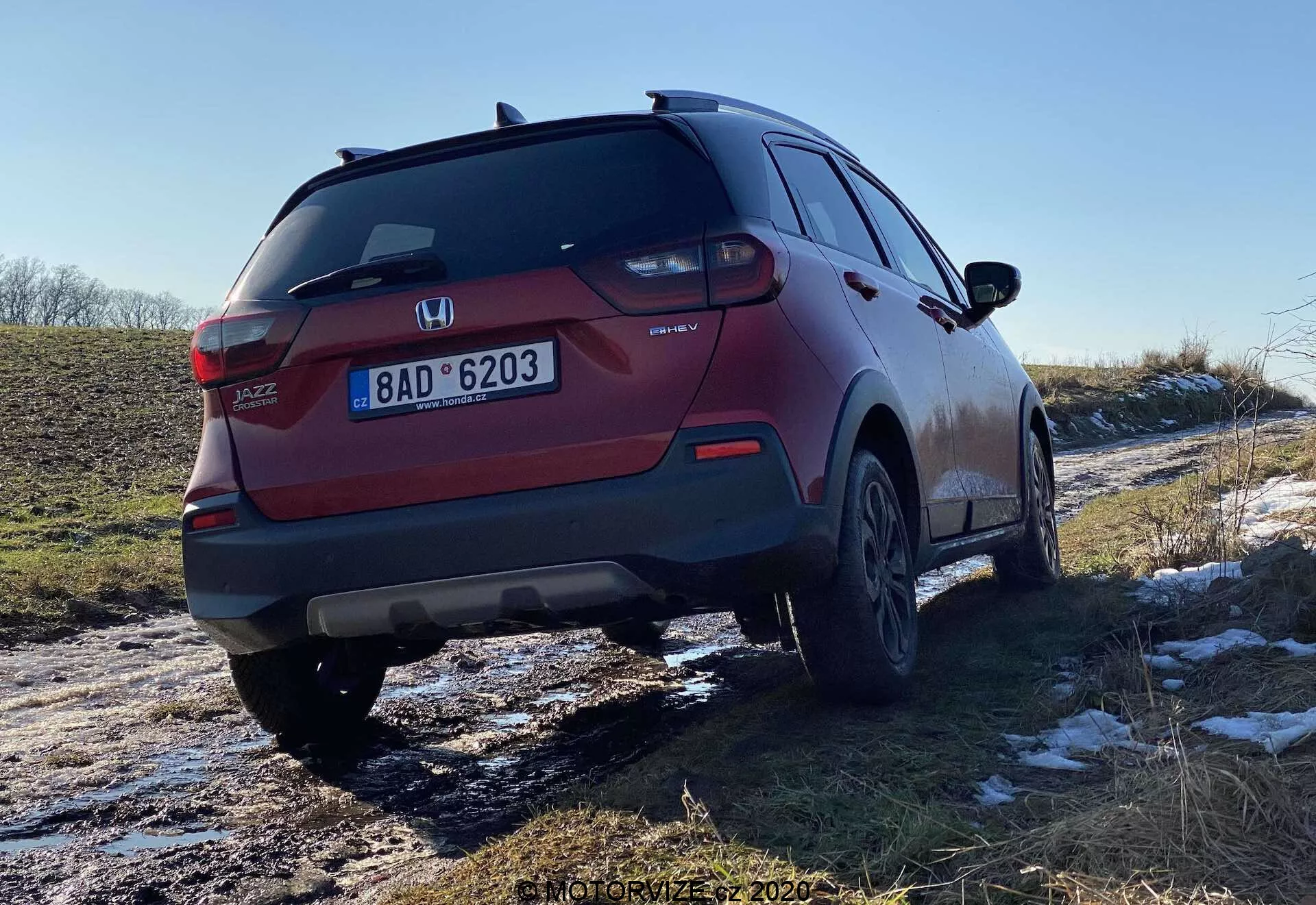 Vista traseira de três quartos de um SUV Honda Jazz Crosstar vermelho, mostrando o design das luzes traseiras, para-choques traseiro, sistema de escape e rodas de liga. O ângulo enfatiza a distância ao solo do veículo e parece ter viajado recentemente em estradas com neve e lama, indicando a sua versatilidade em diversas condições.