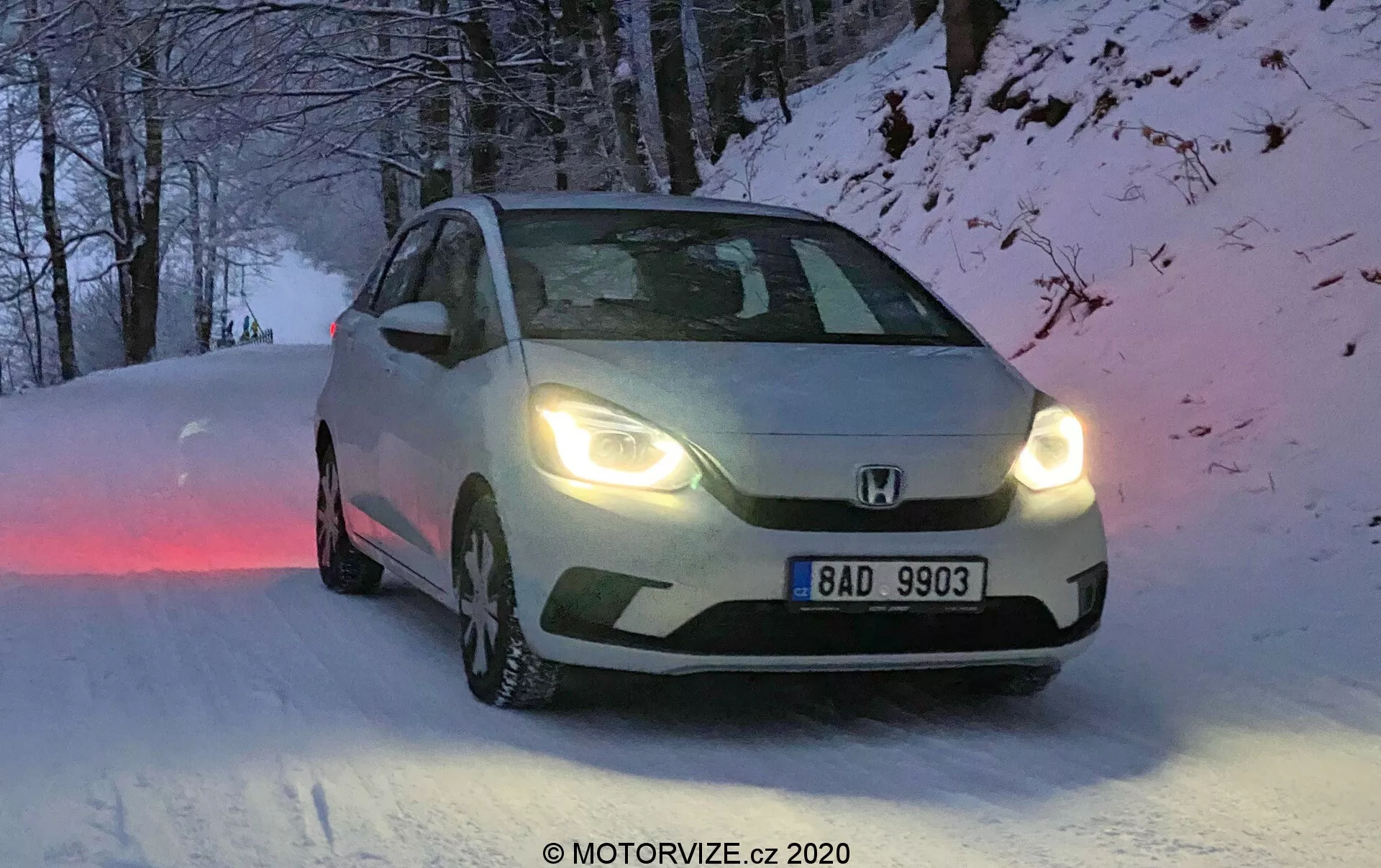 Esta imagem apresenta um Honda Jazz 2020, capturado num ângulo de três quartos da frente, destacando as características de design da grade frontal, faróis e contornos do capô. Posicionado numa estrada coberta de neve, o veículo tem pneus de inverno com padrões de banda de rodagem visíveis e os faróis ligados, indicando condições de amanhecer, anoitecer ou baixa visibilidade. A placa de matrícula europeia diz “BAD 9903”, e a imagem tem um ligeiro desfoque de movimento que adiciona uma qualidade dinâmica. A iluminação natural e fria e o cenário de inverno são complementados pelas capacidades para todo o tipo de tempo do veículo.