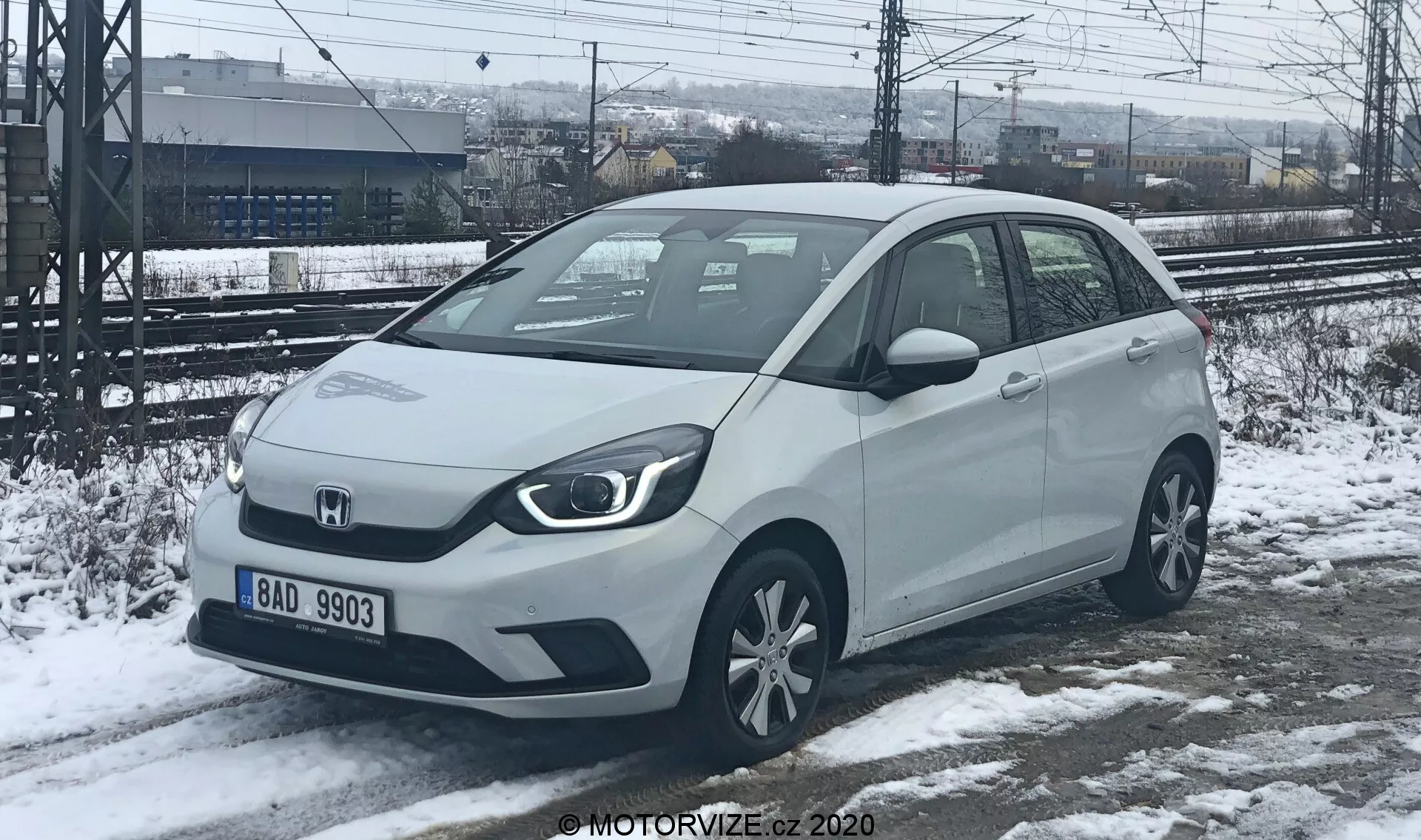 Vista em ângulo de três quartos frontais de um carro compacto Honda Jazz 2020 em uma cor clara, possivelmente branco ou prata, capturado em um cenário de inverno com neve no chão. Os faróis afiados e angulares e a grade frontal sólida sugerem opções de motorização eletrificada. O carro possui sensores de estacionamento visíveis no para-choque dianteiro, espelhos laterais montados nas portas com sinais de direção integrados e rodas de liga leve padrão. Estacionado em neve parcialmente derretida, com trilhos de trem e uma cerca ao fundo, indicando uma localização industrial ou próxima a uma estação ferroviária.