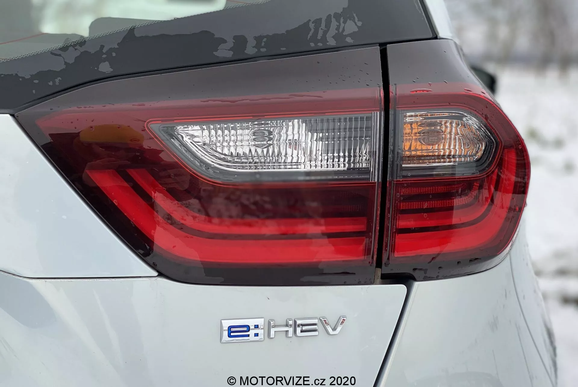 Honda Jazz 2020 Close-Up da Parte Traseira: Com Emblema Híbrido 'e:HEV' e Luz Traseira LED Moderna. A imagem destaca a carroceria cinza-prateada com design intrincado da luz traseira em um ângulo baixo de 45 graus.