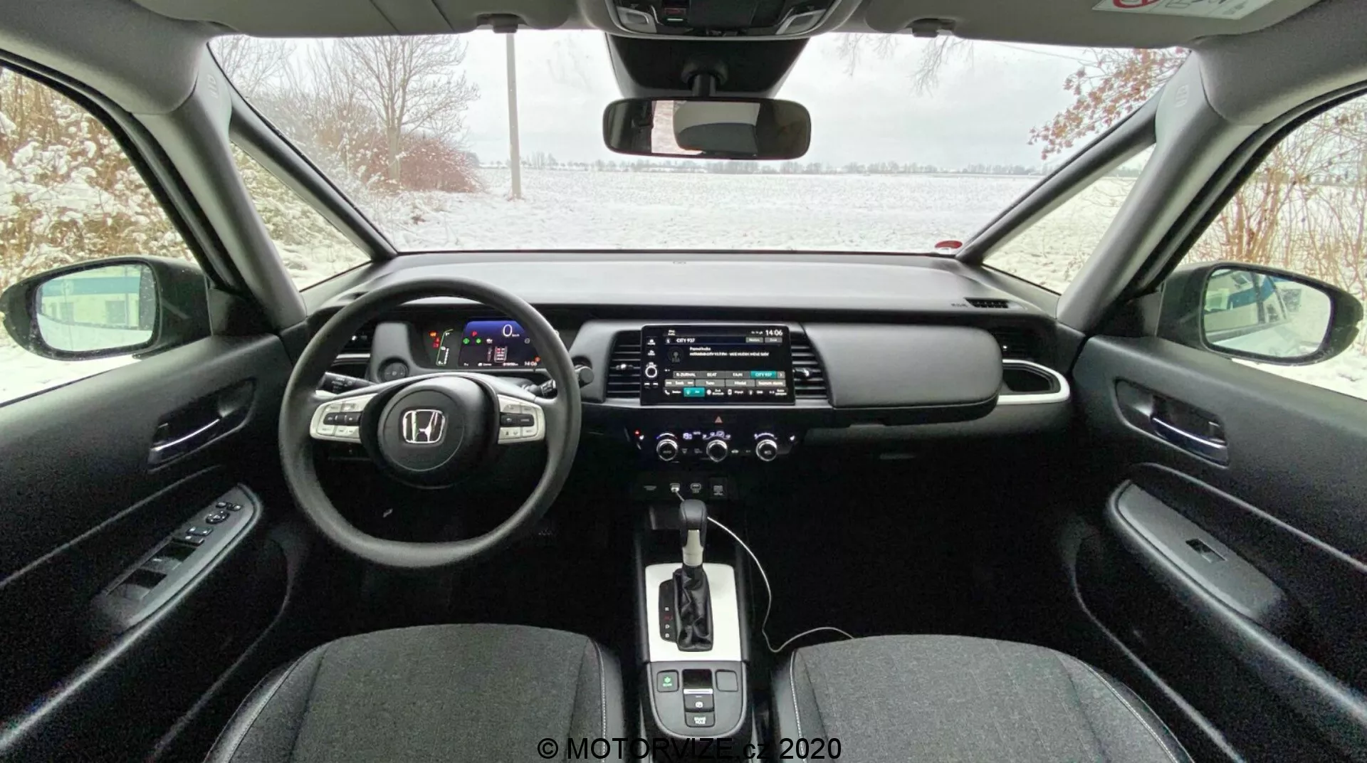 Interior de carro moderno visto do banco traseiro, apresentando painel de instrumentos digital, volante multifuncional, grande tela sensível ao toque do sistema de infoentretenimento, caixa de câmbio automática com alavanca de couro, controle de clima de zona dupla, freio de estacionamento eletrônico e design elegante com materiais contrastantes; exterior nevado visível pelo para-brisa indicando uma configuração possivelmente invernal.