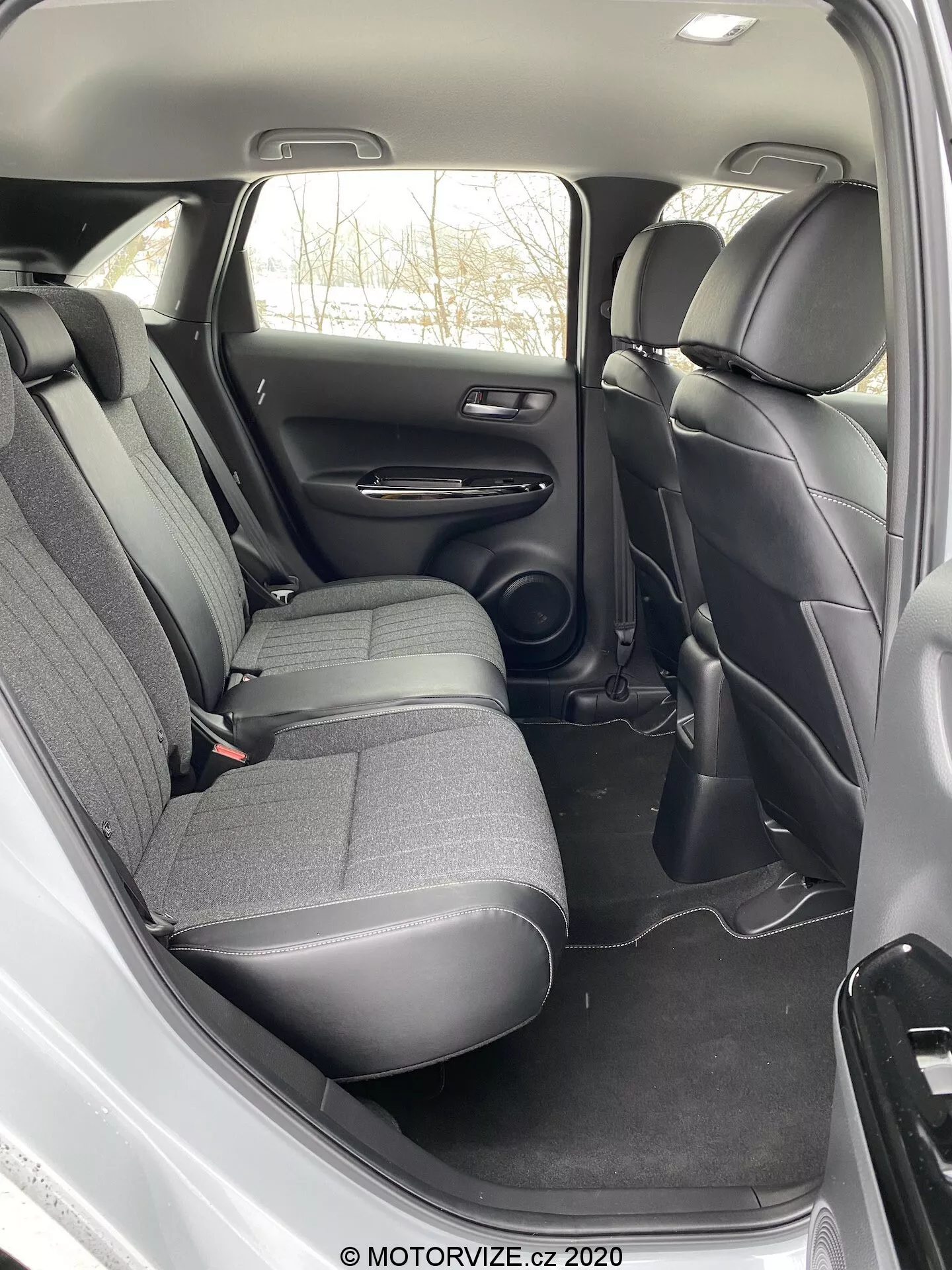 A imagem mostra o compartimento traseiro para passageiros do interior do Honda Jazz 2020. A vista, tirada de um ângulo ligeiramente elevado acima e ao lado da porta traseira aberta, revela um banco traseiro com encostos de cabeça para três passageiros, estofado em um tecido escuro durável. O painel da porta inclui controles, um alto-falante, um descansa-braço e um compartimento de armazenamento, enquanto o chão é coberto por um carpete de tom escuro que combina com o restante. Entre os assentos dianteiros, um console central se estende até a parte traseira, sem controle climático traseiro visível ou pontos de carregamento. As características de segurança visíveis incluem cintos de segurança de três pontos. O design é prático com materiais padrão, enfatizando durabilidade e funcionalidade ao invés de luxo.