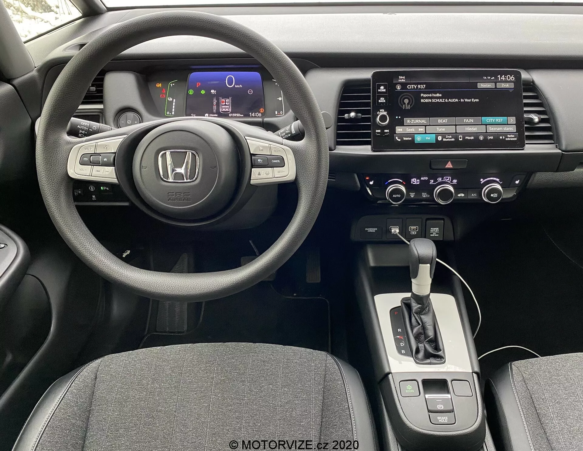 Interior de um Honda Jazz 2020 na perspectiva do motorista, mostrando um volante de três raios com controles montados, um painel de instrumentos digital com medidores coloridos, uma consola central com sistema de infoentretenimento touchscreen, controles manuais de áudio e clima, um seletor de transmissão automática e assentos revestidos em tecido.