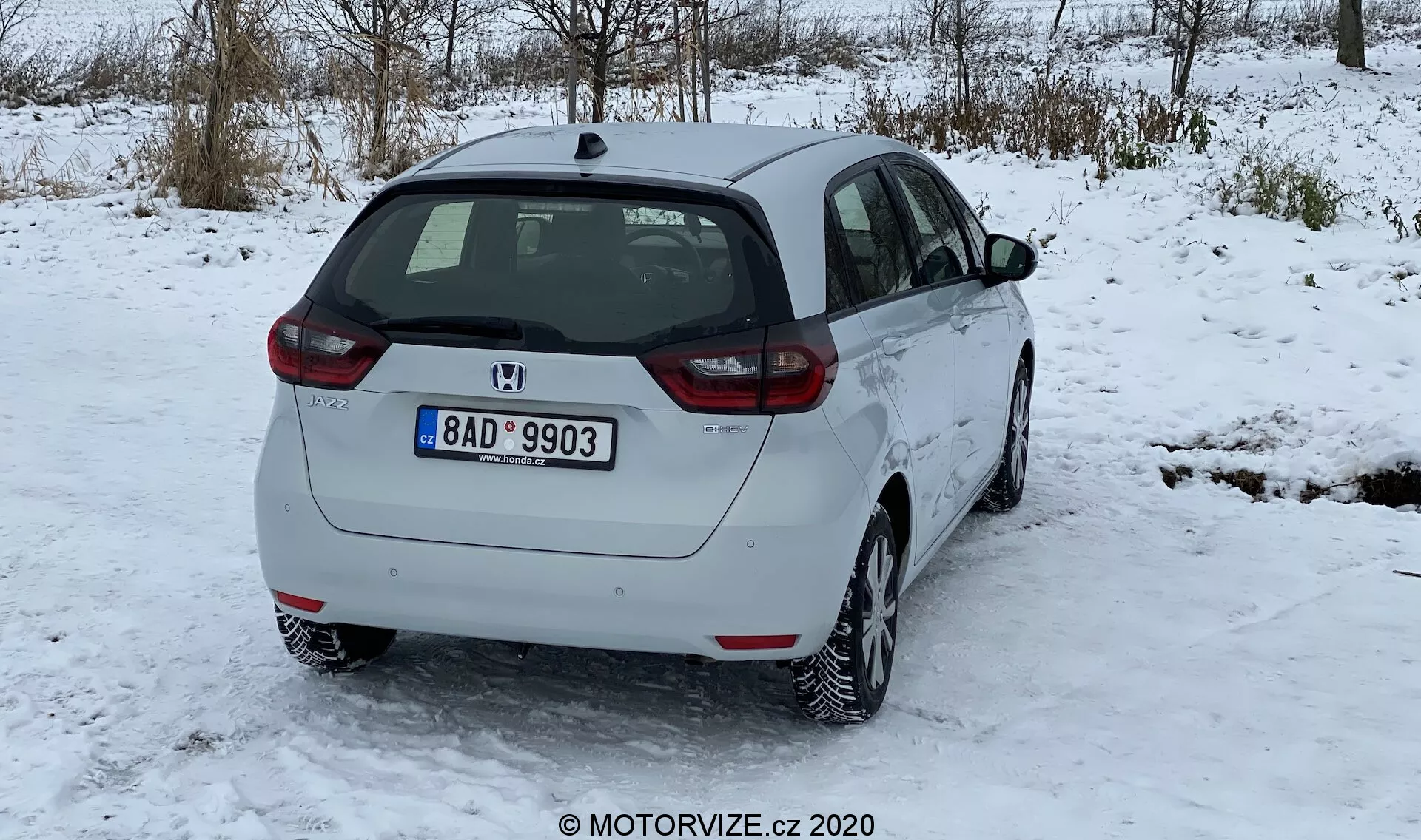 A imagem mostra uma vista traseira de três quartos de um Honda Jazz 2020 branco, estacionado em uma superfície coberta de neve, com árvores sem folhas no fundo de cenário invernal e um céu nublado. As características do design traseiro incluem um limpador de para-brisa traseiro, o emblema do fabricante, a indicação do modelo e o conjunto de iluminação traseira com lentes vermelhas e transparentes. O perfil lateral mostra a porta traseira do passageiro, a porta dianteira, as janelas laterais e um tampão de combustível visível. O carro está equipado com rodas de liga leve de múltiplos raios, indicando um padrão de pneu adequado para neve com acumulação menor de neve. A placa de número traseira sugere sua origem geográfica. A distância ao solo é típica dos hatchbacks, sem ponteira de escape visível ou acessórios exteriores. O carro parece limpo e em boas condições, com pouca sujeira no para-choque traseiro devido a estradas nevadas.