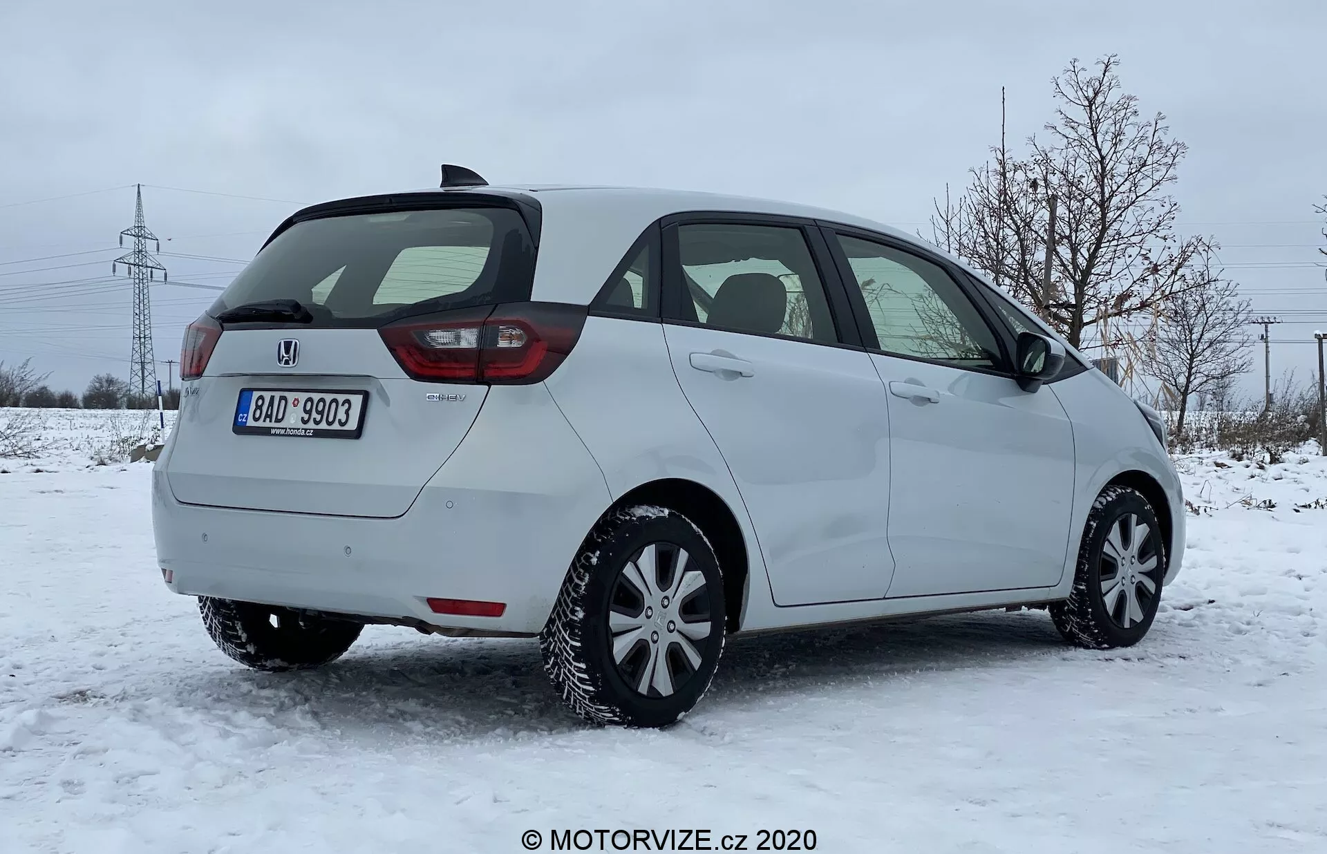 Vista traseira de três quartos de um carro hatchback Honda Jazz 2020, numa superfície coberta de neve, num ambiente suburbano com árvores despidas e torres elétricas ao fundo. A imagem captura o lado do passageiro do carro, design moderno das luzes traseiras provavelmente com tecnologia LED, porta traseira, limpador de pára-brisa traseiro, para-choque traseiro visível com sensores de estacionamento integrados e aparentemente pneus de inverno. O veículo está estacionado em luz natural do dia sob um céu nublado, sugerindo condições de condução de inverno.