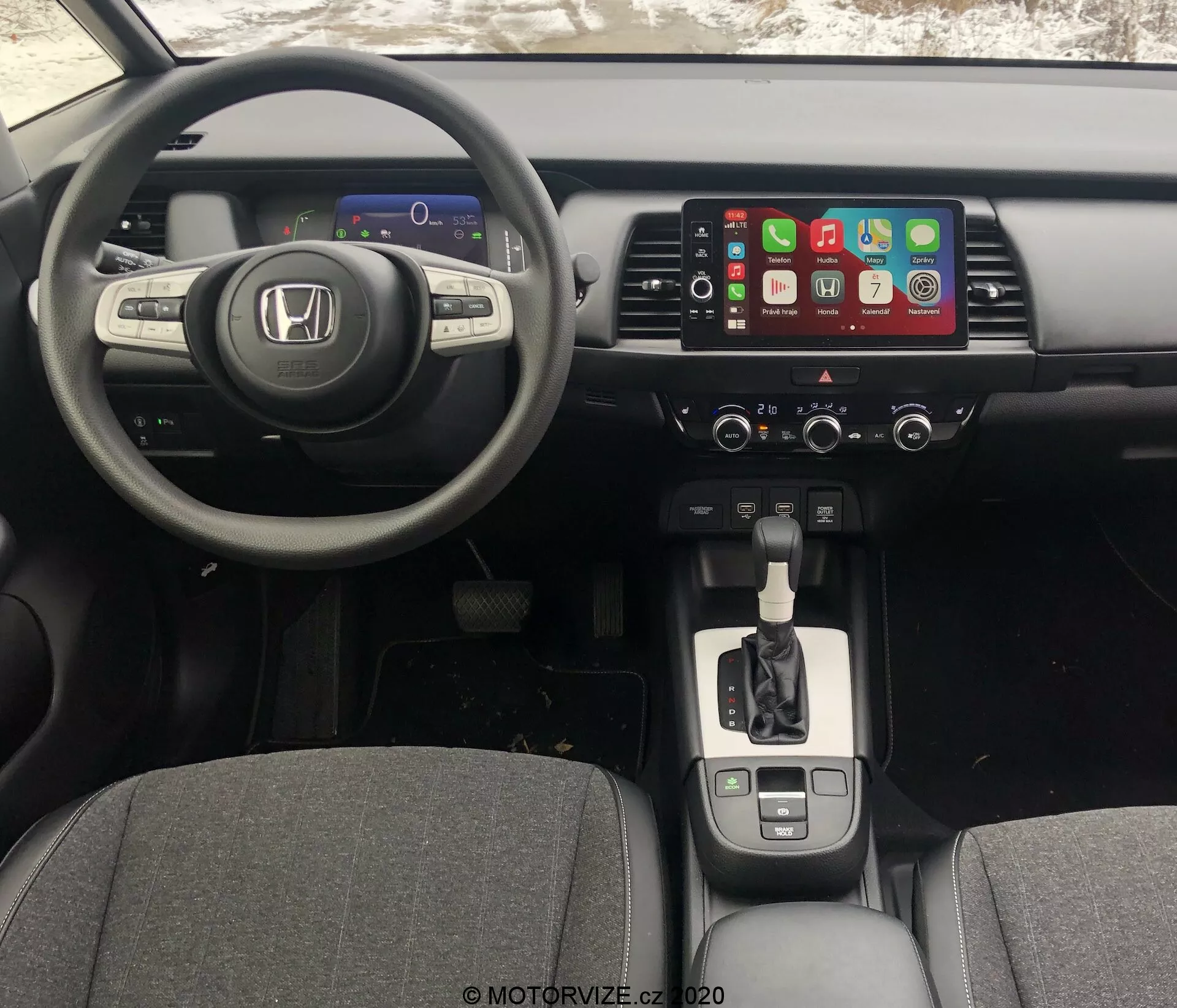 Vista do interior do Honda Jazz 2020 a partir da perspectiva do motorista, mostrando o painel, o volante e o console central. O ângulo ligeiramente elevado revela o painel de instrumentos digital, o volante multifuncional de três raios, o sistema de infotainment com tela sensível ao toque compatível com Apple CarPlay, os controles de clima, a alavanca de câmbio com acabamento cromado, os porta-copos, o freio de estacionamento eletrônico e a conectividade USB.