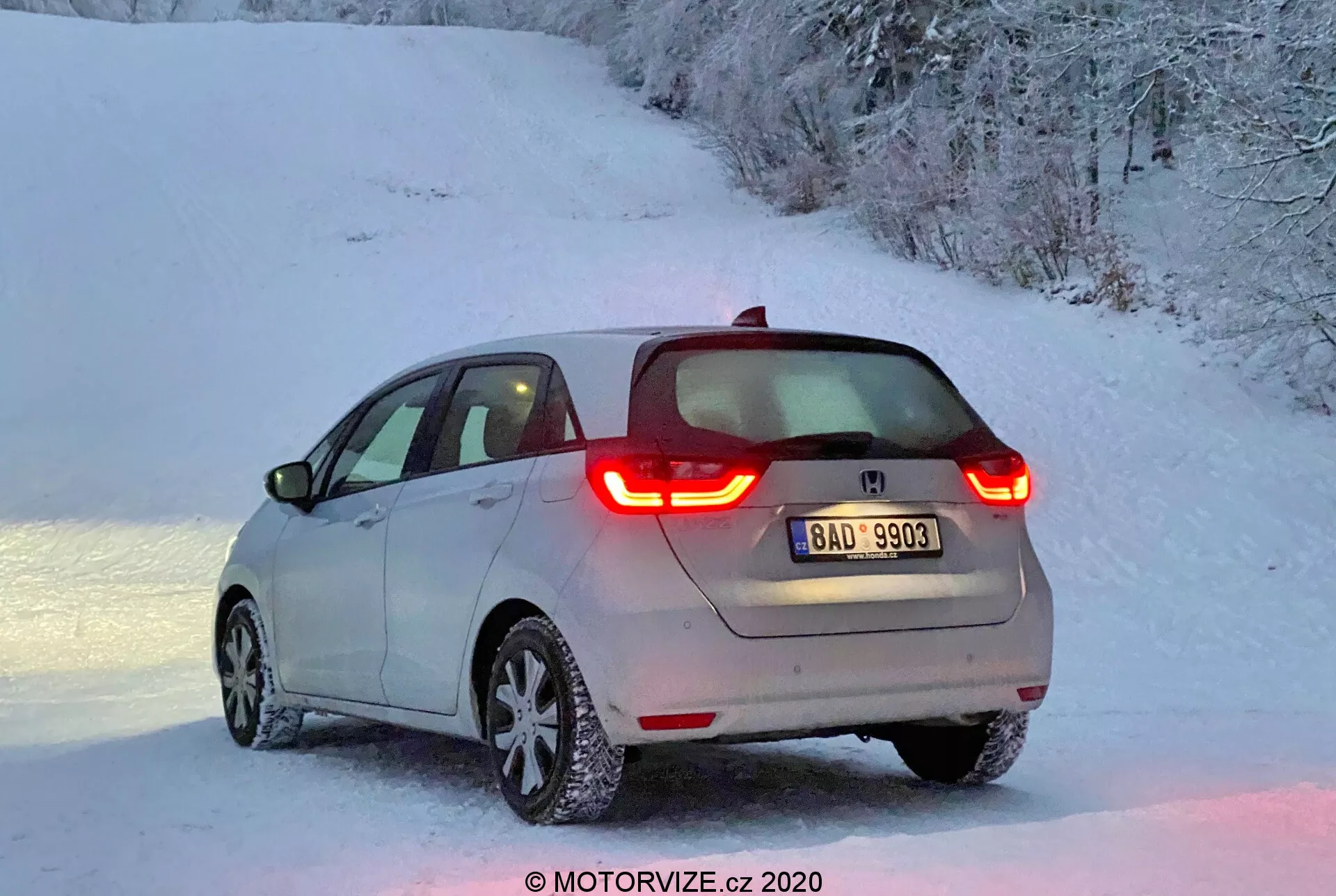 Vista traseira três quartos de um moderno Honda Jazz 2020 hatchback, iluminado pela suave luz do amanhecer ou do entardecer, com luzes traseiras de LED em forma de C iluminadas, um logotipo do fabricante visível, uma leve camada de neve no veículo e rodas de liga leve com pneus cobertos de neve em um cenário rural nevado.