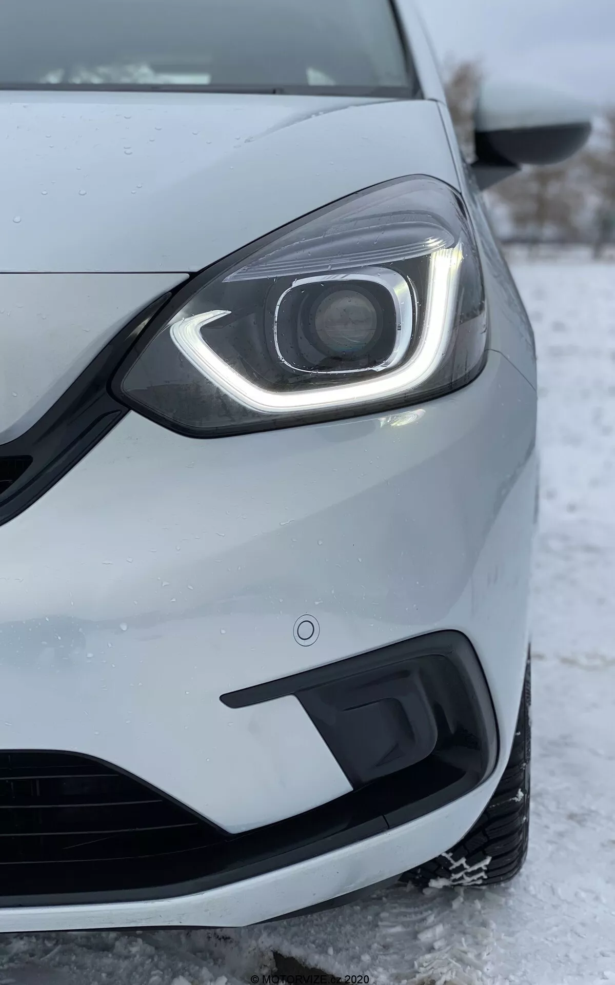 Imagem em close-up do lado frontal esquerdo de um Honda Jazz 2020, tirada de um ângulo baixo. O foco está no farol que apresenta um design moderno em LED com luzes diurnas que curvam ao redor da parte superior e lateral. O conjunto do farol inclui uma lente de projetor visível, sugerindo faróis HID ou LED avançados para o feixe principal. O para-choque dianteiro, mostrado abaixo do farol, inclui uma grade de entrada de ar e uma carcaça para luzes de neblina. A pintura metálica prateada ou cinza claro do carro e a roda parcialmente visível e o espelho lateral são detalhados. Neve e gelo no chão indicam um clima frio, refletindo o desempenho do carro em condições de inverno. A imagem também destaca a qualidade de construção do carro, os espaços entre os painéis e o sensor de proximidade no para-choque, que faz parte da tecnologia de assistência de estacionamento.