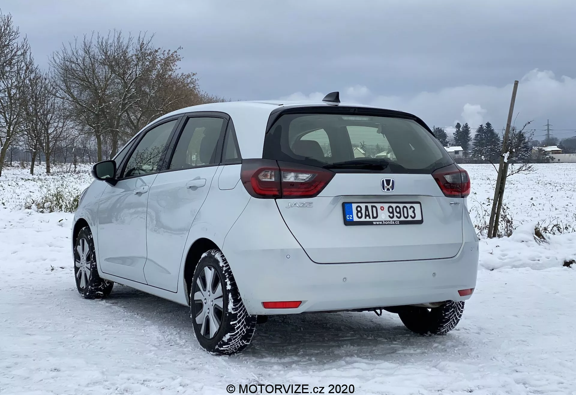 Imagem de um Honda Jazz 2020 prateado estacionado em condições de neve com um cenário rural ou semi-rural ao fundo. Capturado da parte traseira e perfil lateral parcial do lado direito, destacando as luzes traseiras dispostas verticalmente, o emblema central e o para-choques com sensores de estacionamento. Contornos laterais lisos, rodas de liga leve e pneus de inverno são claramente visíveis, junto com a capacidade de desembaçamento nas janelas e no para-brisa traseiro, e espelhos laterais não retráteis da mesma cor da carroçaria. A placa, a antena no teto e o estado geral bem conservado do veículo também são notados.