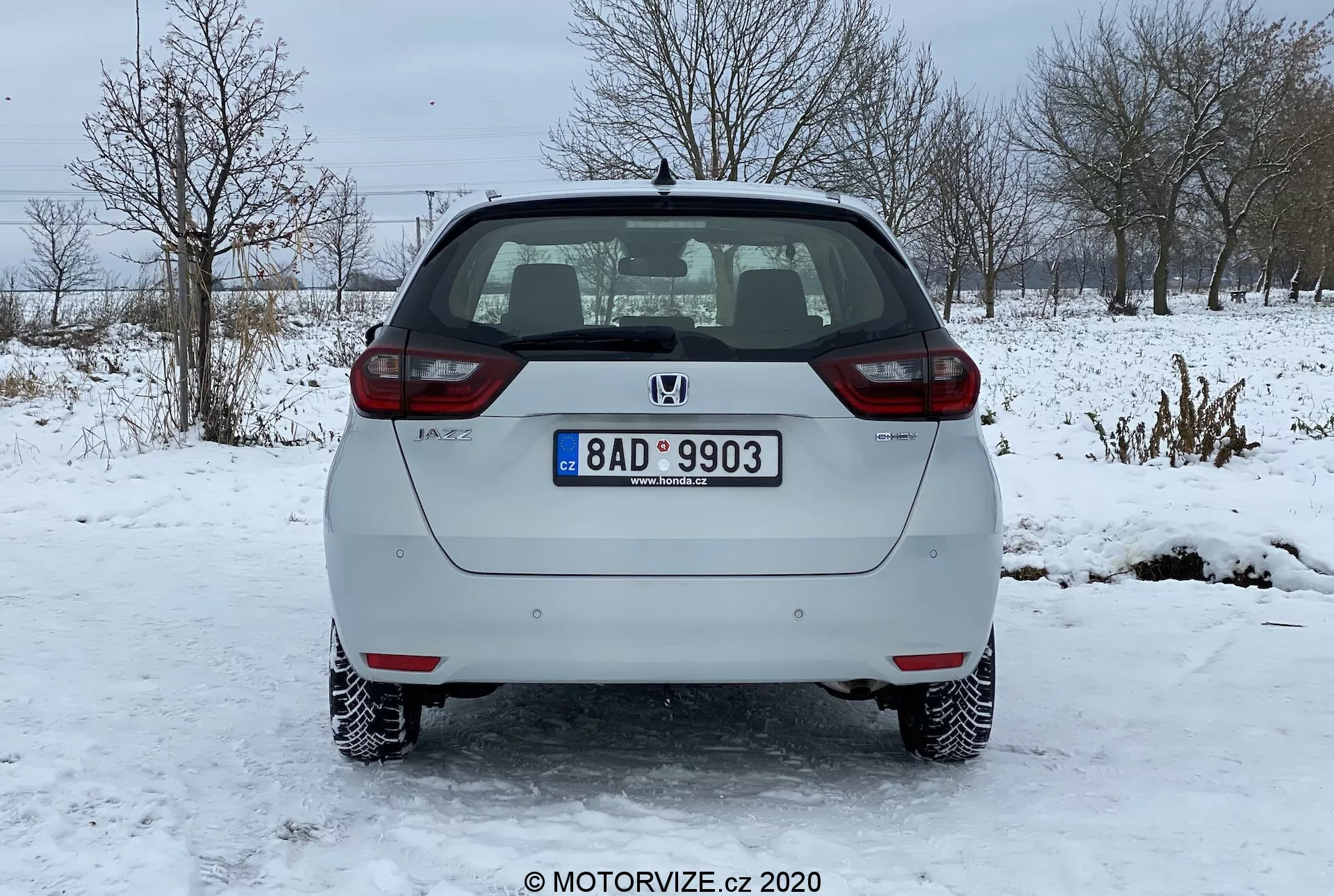 Vista traseira de um carro hatchback prateado Honda Jazz 2020 estacionado em uma superfície gelada. A imagem captura claramente o para-choque traseiro, a porta traseira, as luzes traseiras e o vidro traseiro do veículo. O emblema do fabricante e o nome do modelo são visíveis na porta traseira, junto com uma placa de número padrão montada centralmente. O carro possui luzes de placa iluminadas, refletores em ambas as extremidades do para-choque traseiro e luzes traseiras com lâmpadas padrão. O vidro traseiro tem uma função de descongelamento mostrada por linhas horizontais, e há um limpador de para-brisa traseiro estacionado no topo. O sistema de escapamento é ligeiramente visível abaixo do para-choque traseiro do lado direito. O carro tem rodas de aço com coberturas decorativas e está situado em uma paisagem de inverno com neve e árvores sem folhas ao fundo.