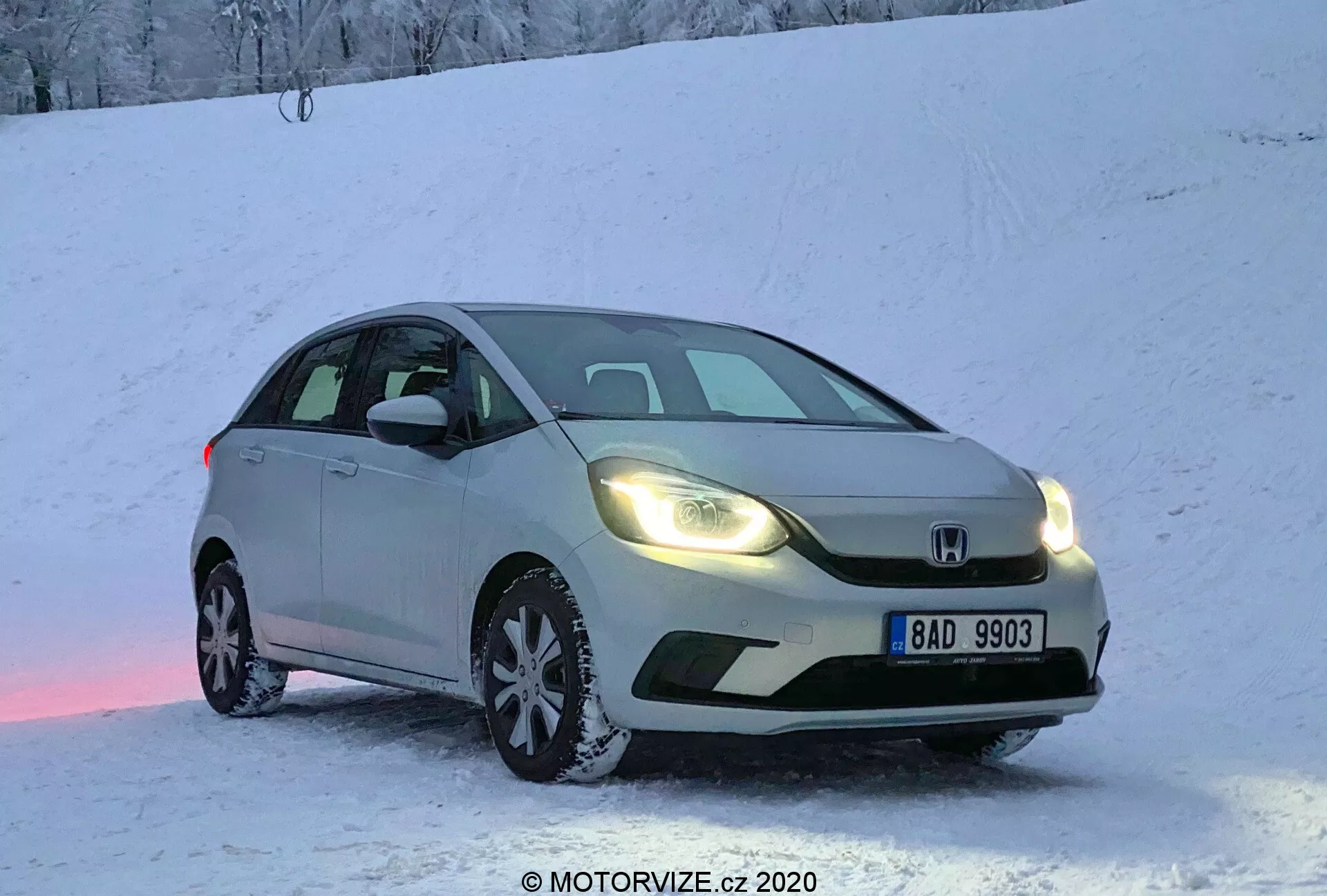 Carro hatchback Honda Jazz 2020 de cor prata estacionado em uma superfície nevada durante o amanhecer ou anoitecer, com os faróis quentes acesos, capturado de uma vista frontal de três quartos. A imagem destaca a grade frontal, faróis, capô, para-brisa e parte do lado do motorista do veículo. Placa europeia visível, neve no chão e no carro, criando um ambiente frio, mas acolhedor. Reflexo de luz rosado na neve sob o carro. Tiro de ângulo baixo que enfatiza o equipamento de pneus de inverno e a aparência dinâmica para capacidades de direção no inverno.