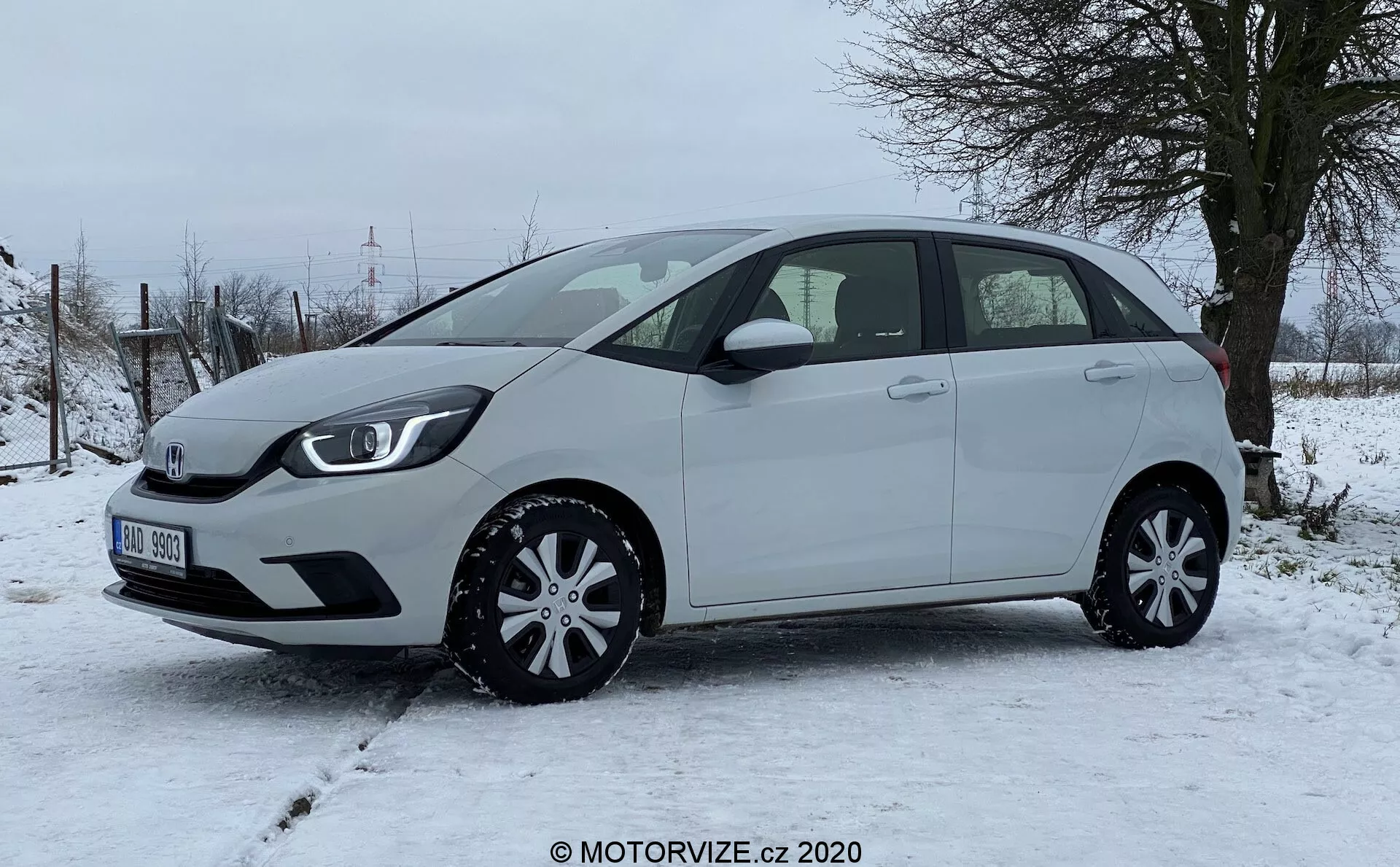 Um moderno Honda Jazz 2020 hatchback fotografado de lado, destacando o perfil esquerdo do veículo em um terreno nevado com vegetação rural ao fundo. O carro compacto apresenta uma linha de teto ligeiramente inclinada, cinco portas, incluindo uma traseira, espelhos laterais da cor do veículo e coberturas de rodas elegantes, exalando praticidade e eficiência no design. Nenhuma característica externa avançada, como câmeras ou sensores, é visível deste ângulo, enfatizando uma visão clara das proporções do carro, contornos das portas, linha das janelas e arcos das rodas.