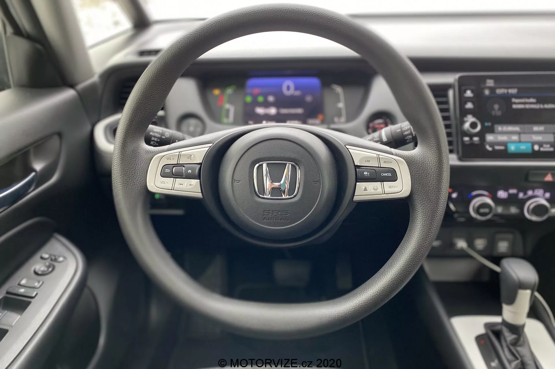 Vista do interior do Honda Jazz 2020 a partir da perspectiva do motorista, destacando o volante de três raios centralizado com emblema Honda, botões de controle para sistema de mídia e controle de cruzeiro nos raios, painel de instrumentos digital ligeiramente desfocado no fundo e console central com sistema de infoentretenimento touchscreen, controles de clima dual zone e alavanca de câmbio automática. Design moderno e contemporâneo com materiais de texturas mistas.