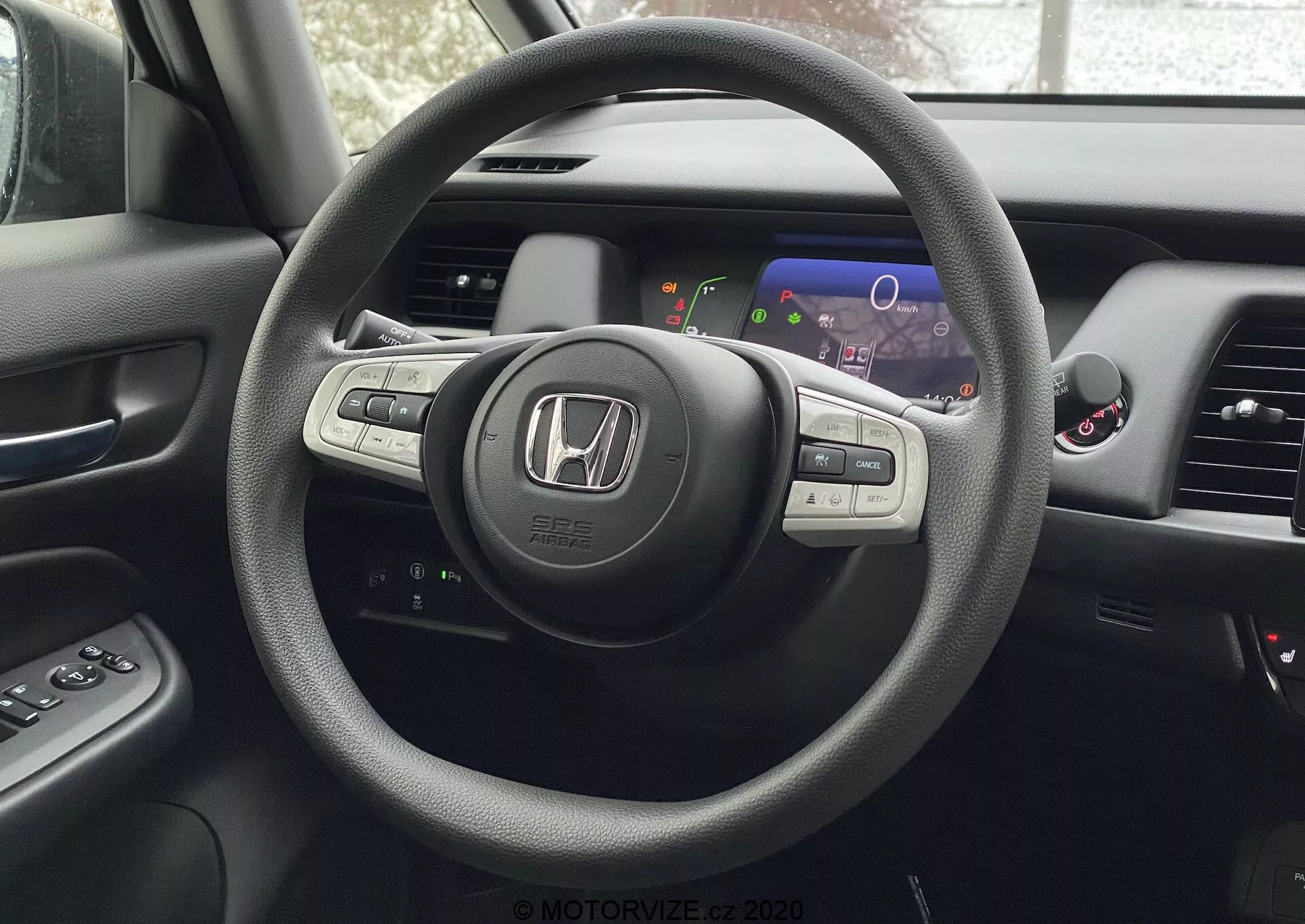 Vista do interior de um Honda Jazz 2020 a partir da perspetiva do banco do condutor, destacando o volante de três raios com emblema Honda e controlos montados para áudio, controlo de cruzeiro e possivelmente Bluetooth ou controlo por voz. Atrás do volante está um painel de instrumentos digital com mostradores coloridos, incluindo velocímetro e tacómetro. Parcialmente visíveis estão o console central com ecrã de infotainment ou navegação e saídas de ventilação. O painel da porta do condutor com botões de controlo das janelas e espelhos laterais também é parcialmente visível.