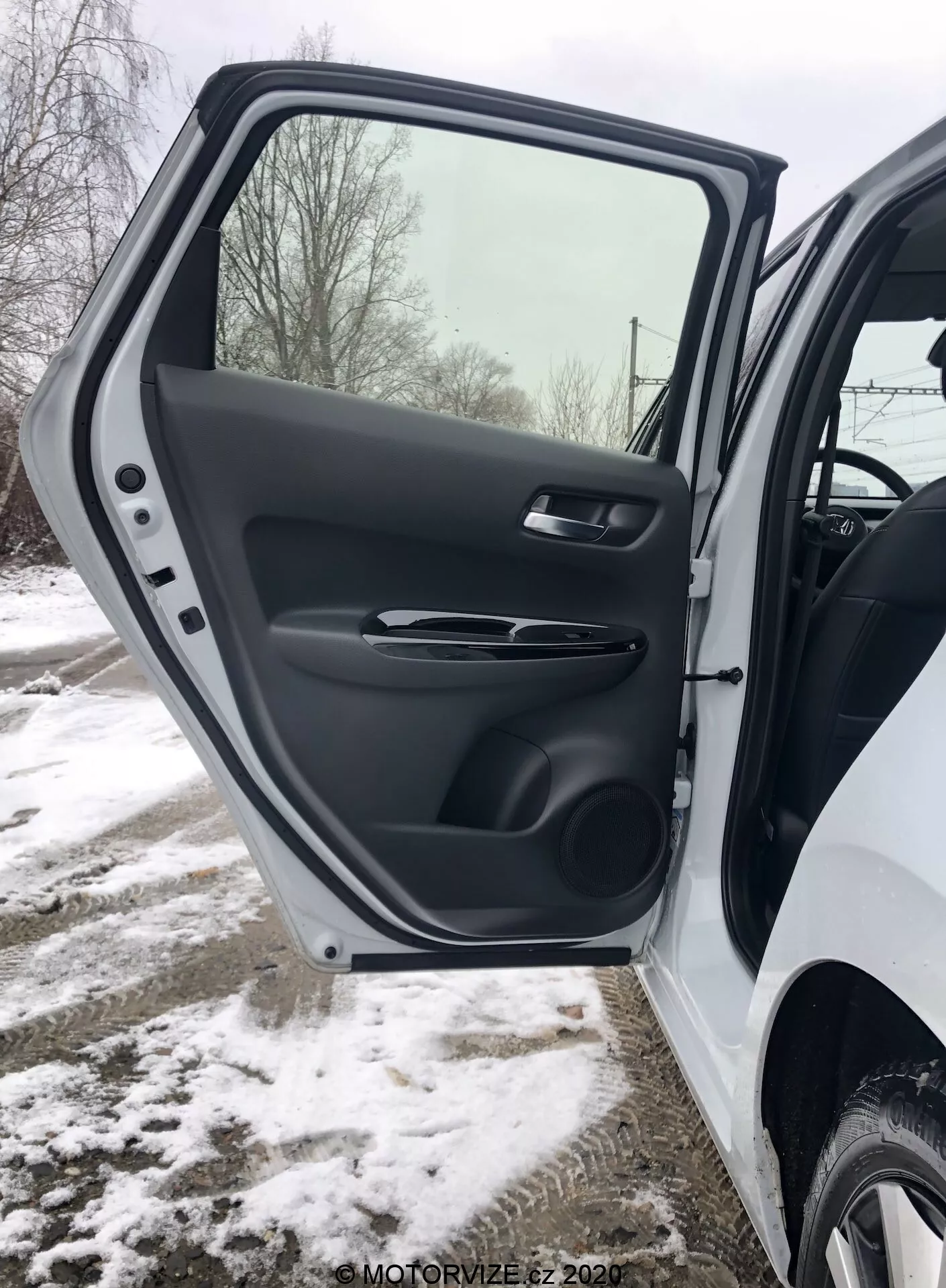 Close-up do interior da porta dianteira do lado do condutor do Honda Jazz 2020, mostrando uma visão detalhada do acabamento em plástico texturizado, maçaneta de porta ergonômica com acabamento acetinado, controles de janelas elétricas e espelhos, bolso de armazenamento moldado e grade do alto-falante, tudo ambientado em um fundo de inverno nevado.